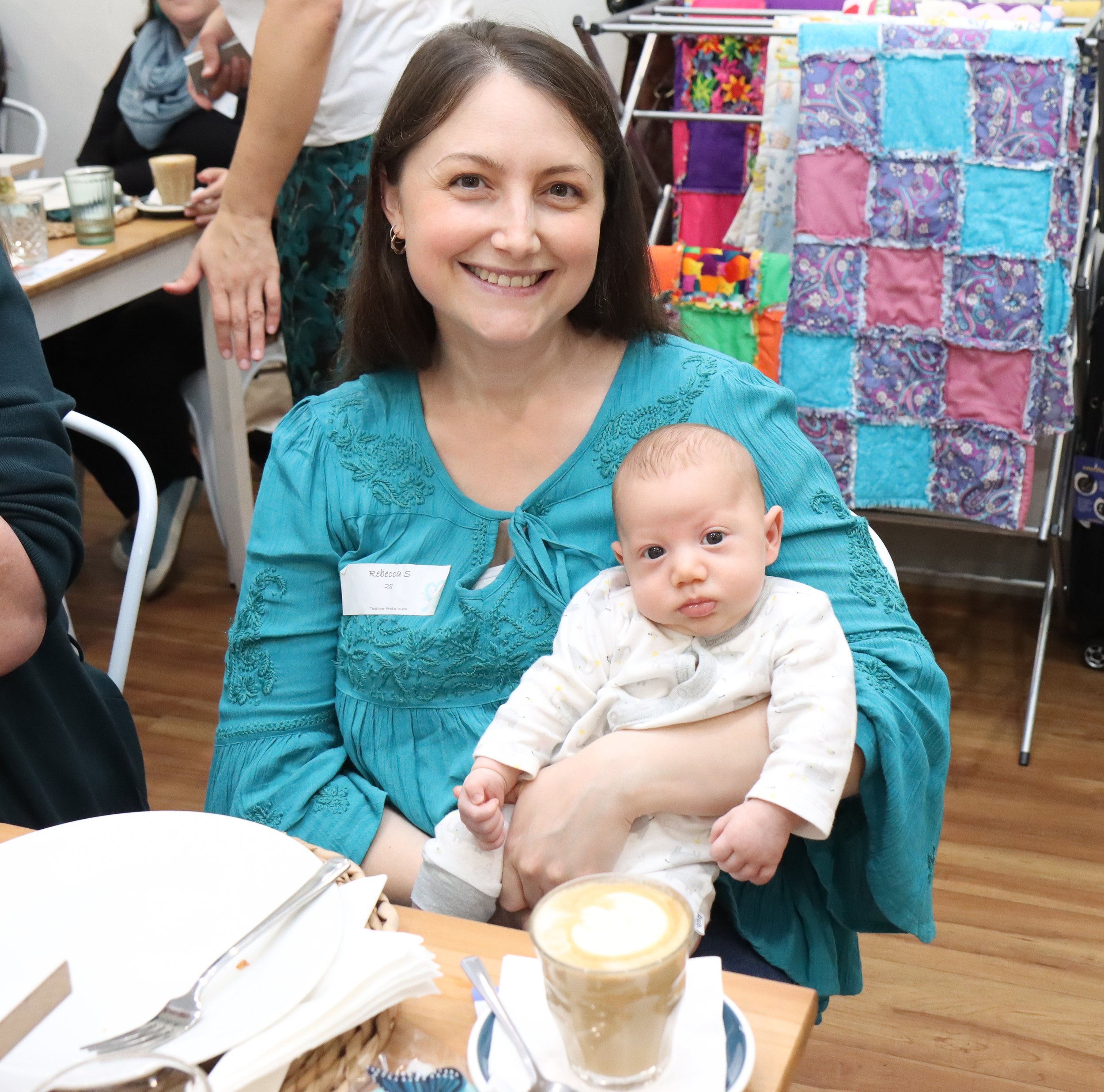 A woman with brown hair wearing a teal embroidered blouse is smiling at the camera while holding a baby with light hair, dressed in a white pajama. They are sitting at a table with a latte and some silverware in front of them, in a room with colorful