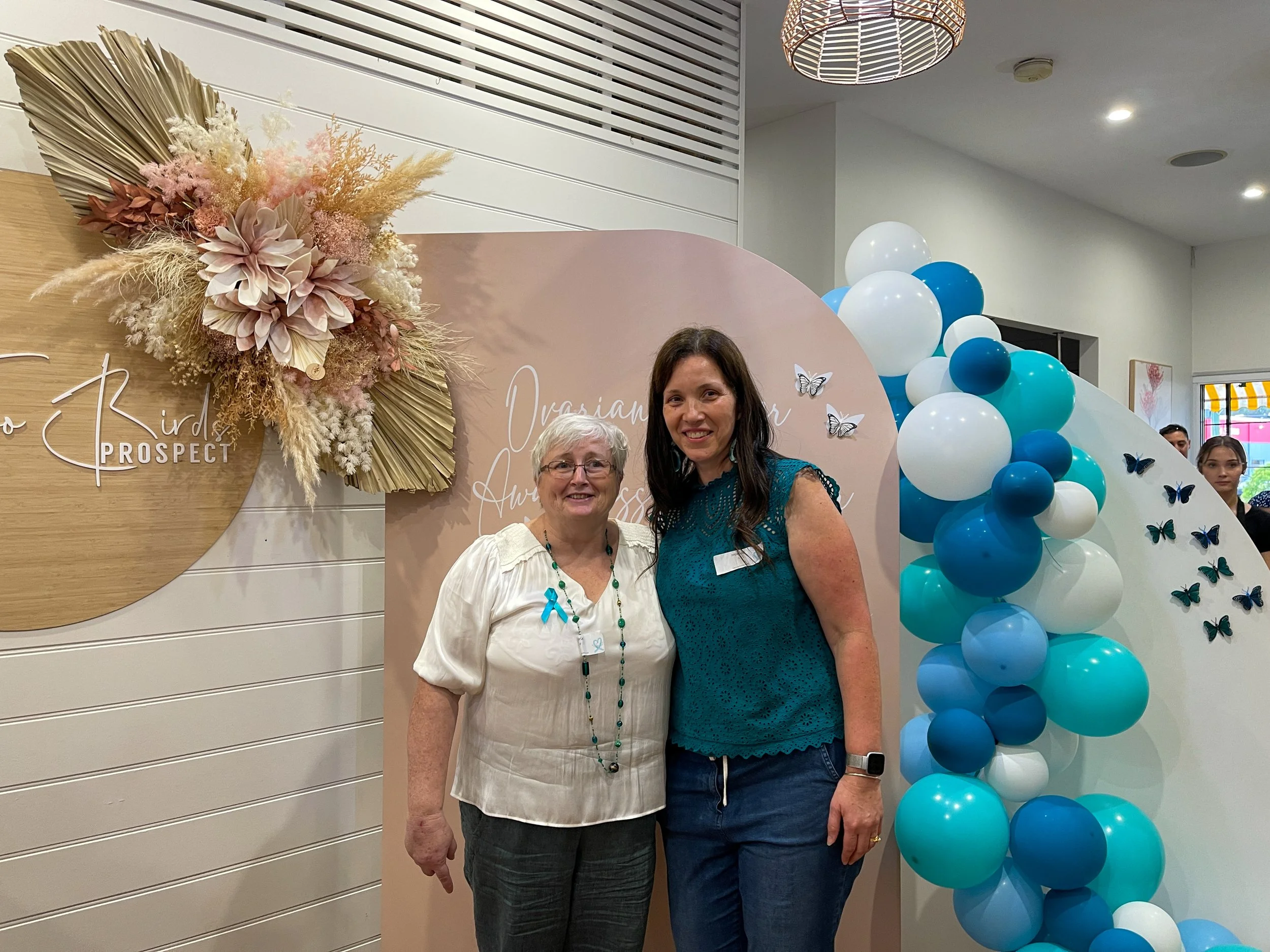 Two women standing next to each other at an event, smiling, with decorative balloons and flowers in the background.