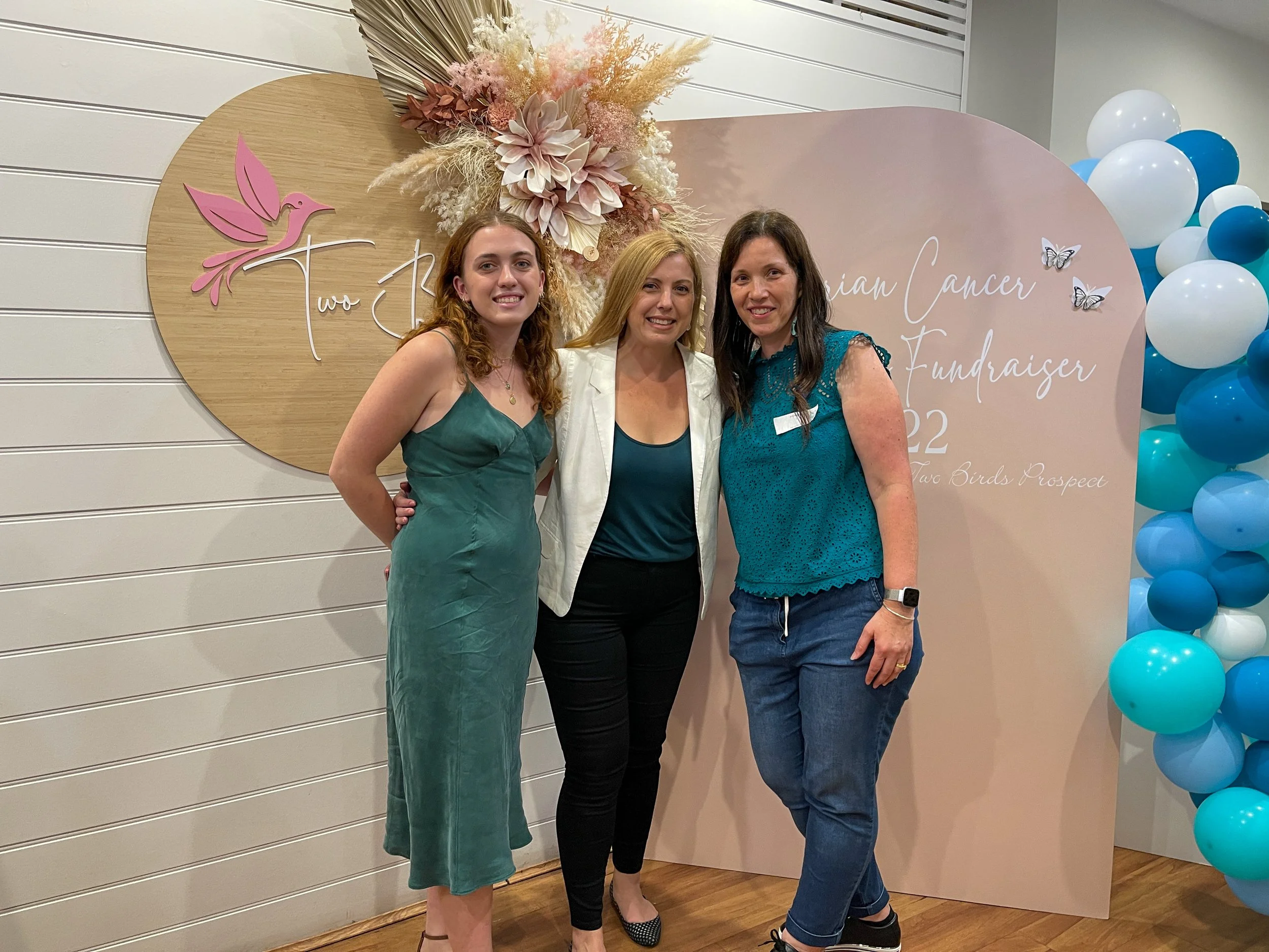 Three women standing close together, smiling, at a breast cancer fundraiser event with pink, white, and blue balloons and a decorative backdrop featuring a pink bird and flowers.