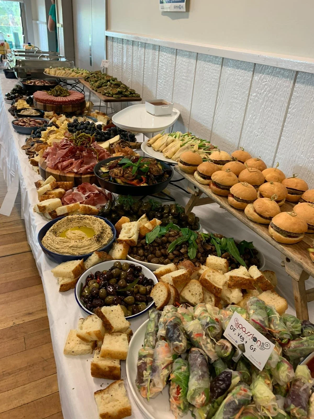 A long buffet table with an assortment of appetizers, salads, sandwiches, olives, hummus, spring rolls, and small burgers in a brightly lit room.