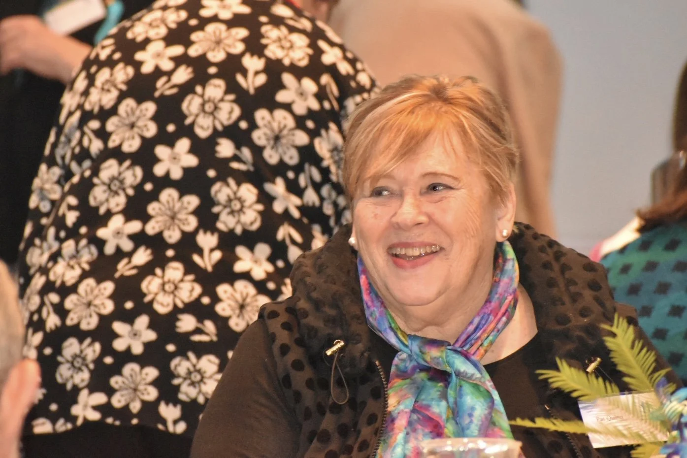 A smiling middle-aged woman with short, light brown hair, wearing a colorful scarf and earrings, in a lively gathering setting.