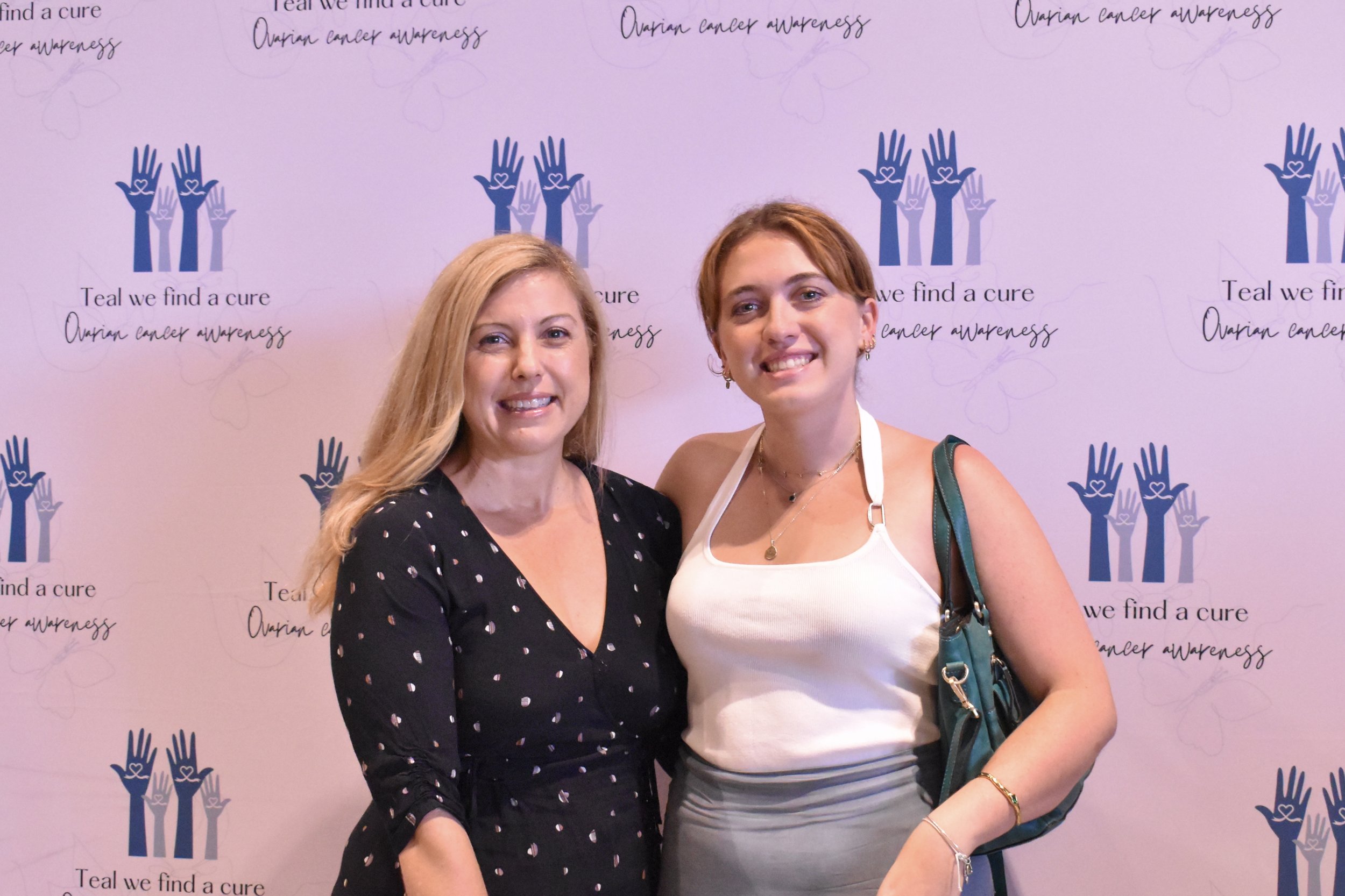 Two women standing together smiling at an event for ovarian cancer awareness, with a pink backdrop featuring the logo and text for 'Teal we find a cure' and 'Ovarian cancer awareness'.