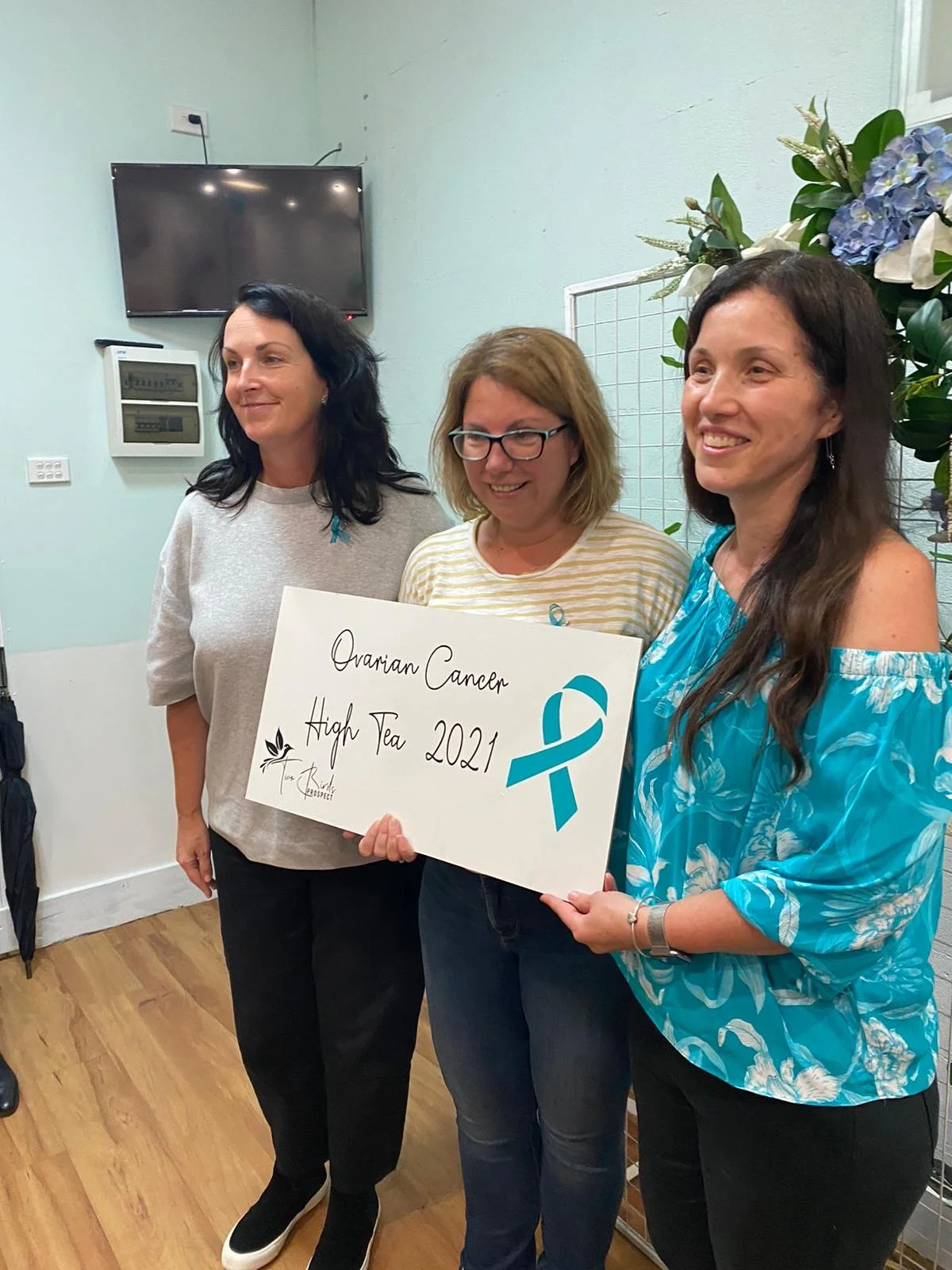 Three women standing indoors holding a sign that says "Ovarian Cancer High Tea 2021" with a teal ribbon symbol. The woman on the right is wearing a blue patterned blouse, the woman in the middle has glasses and a striped shirt, and the woman on the l