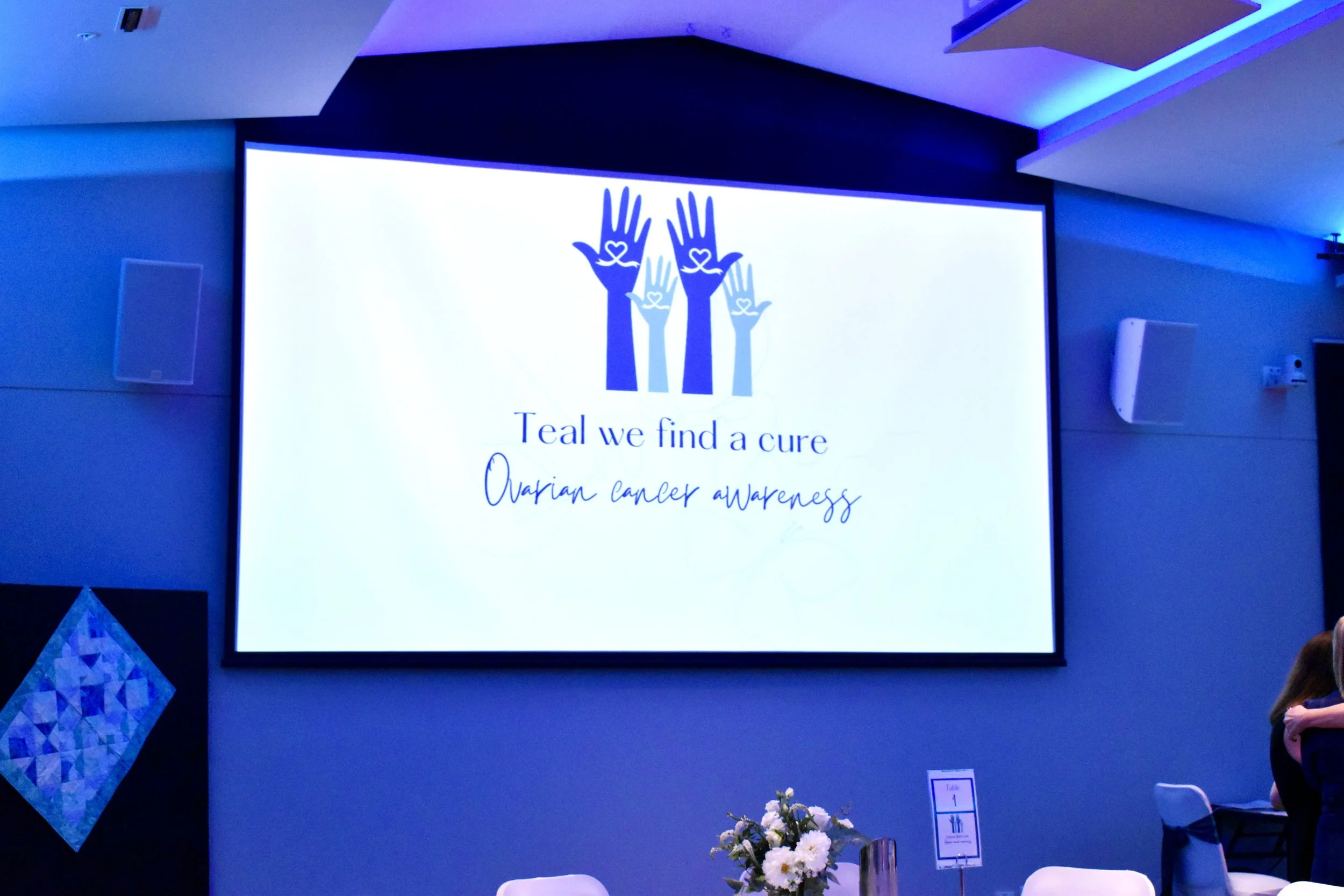 A large screen displays a logo with three stylized hands and text that reads 'Teal we find a cure, Ovarian cancer awareness' at an event.