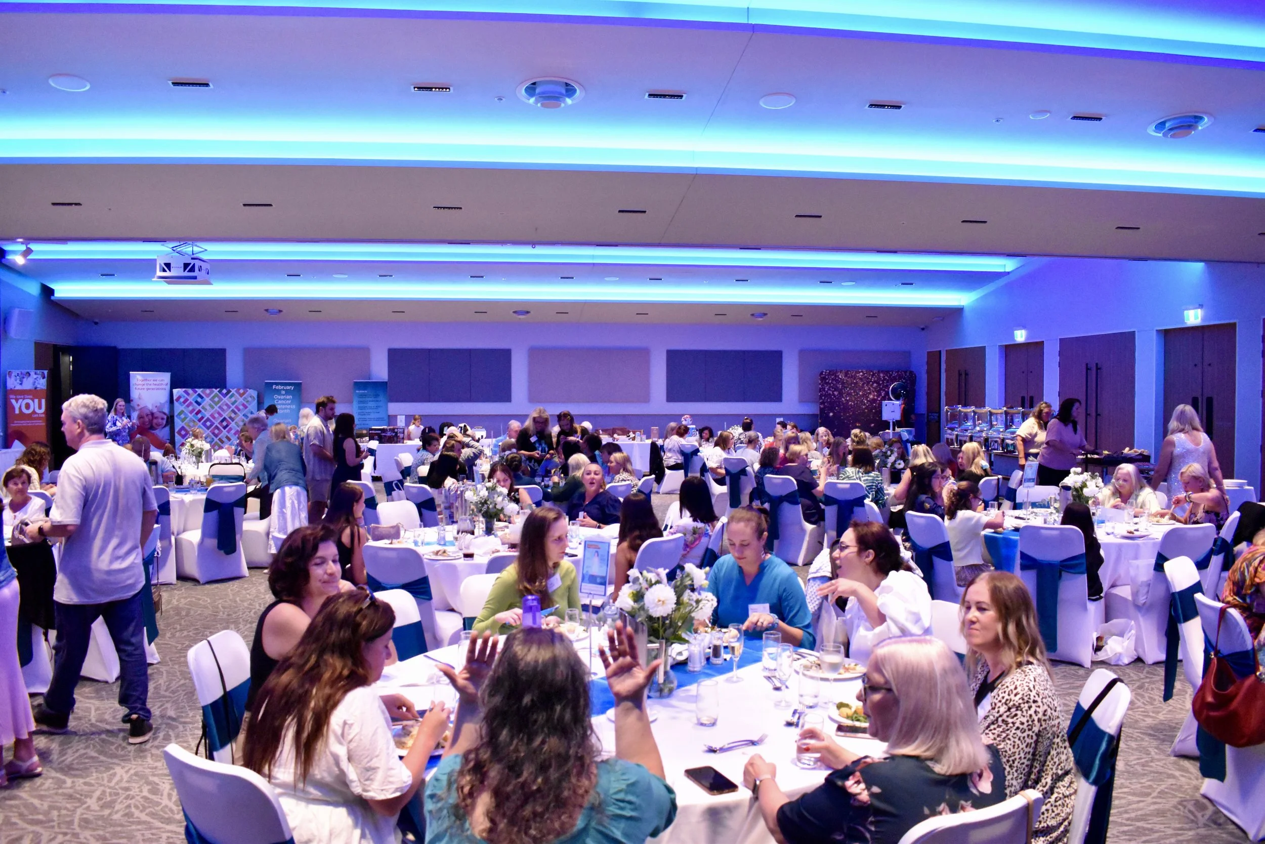 A large banquet hall filled with round tables covered in white tablecloths and decorated with white floral centerpieces. Numerous people are seated, engaging in conversation and enjoying a meal. The room has blue LED lighting on the ceiling and several informational banners and food stations along the sides.