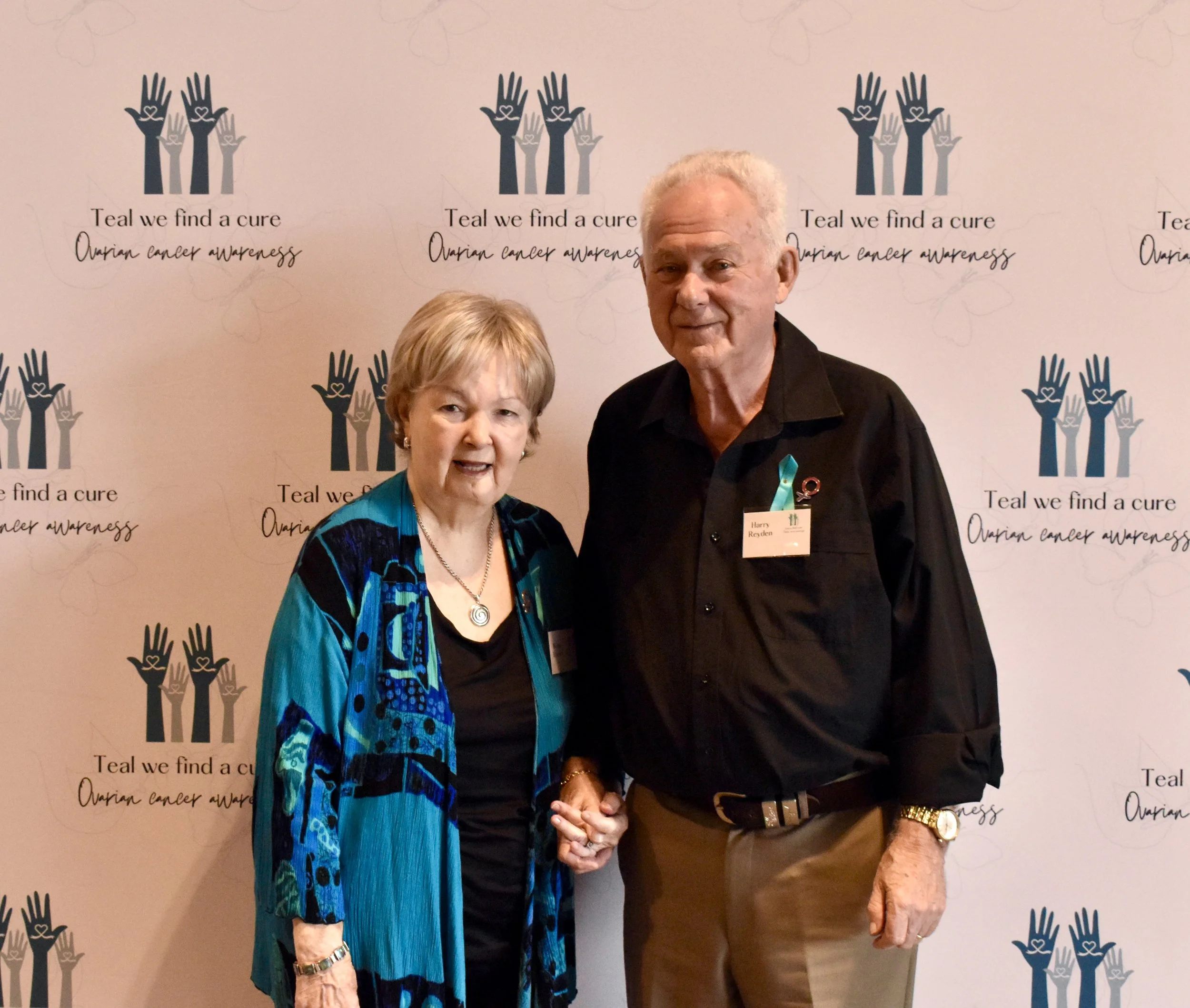 An elderly woman and man standing together in front of a backdrop with ovarian cancer awareness logos, holding hands and smiling.
