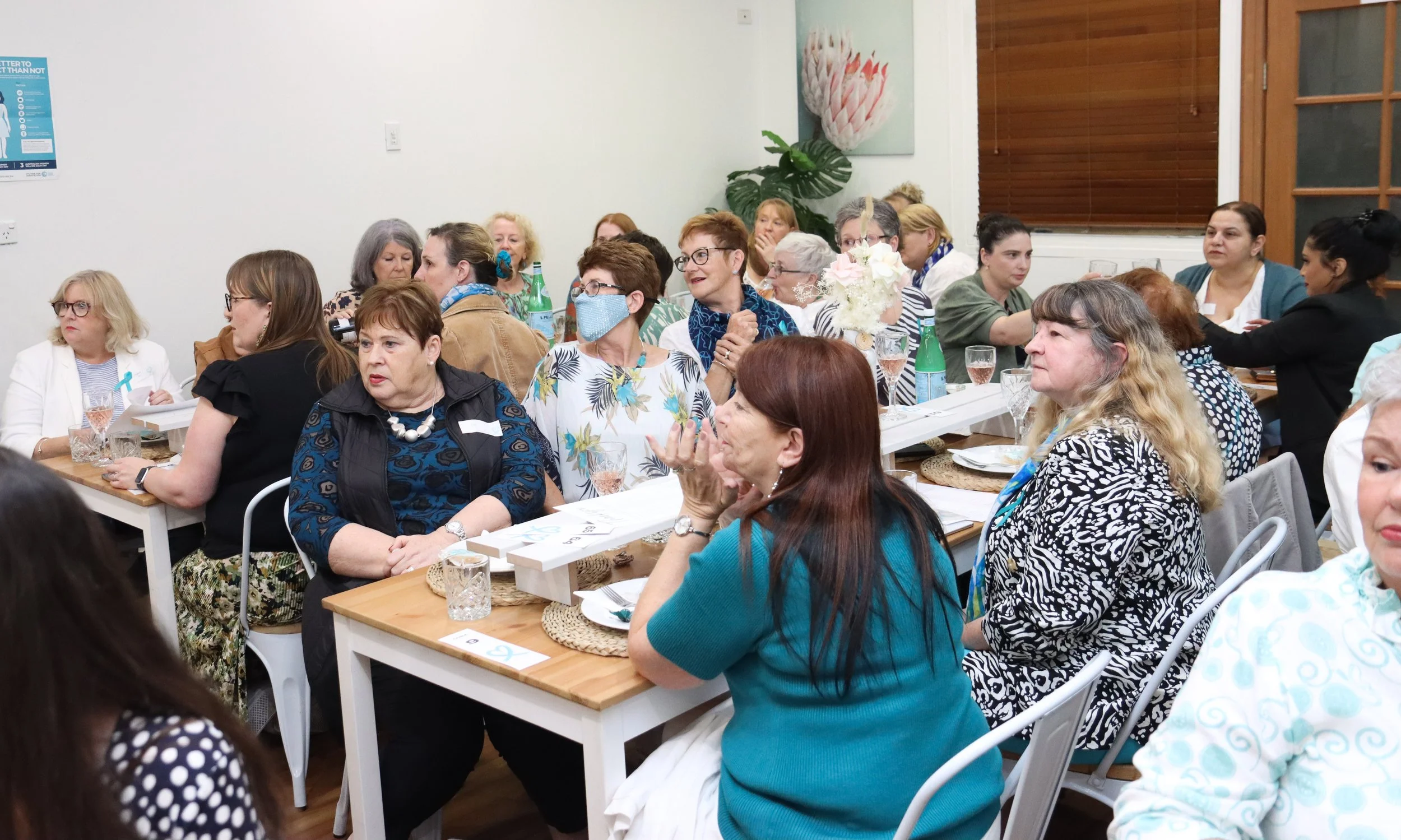 A group of women gathered at long tables in a bright room, attending a meeting or event. They are engaged, some are talking, and there are glasses and bottles on the tables.