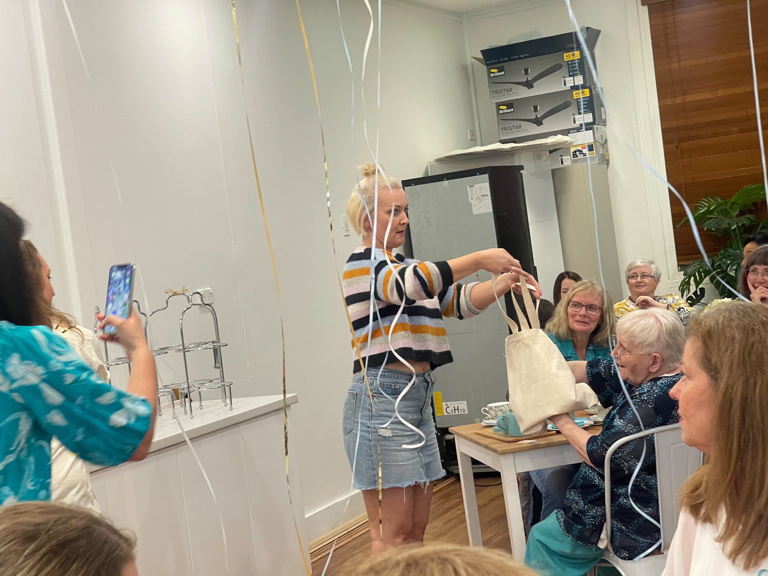 A woman in a striped sweater and denim shorts is handing a gift bag to an elderly woman with white hair, sitting at a table during a celebration. Several other older women are seated at the table, smiling and watching the exchange, with some taking p