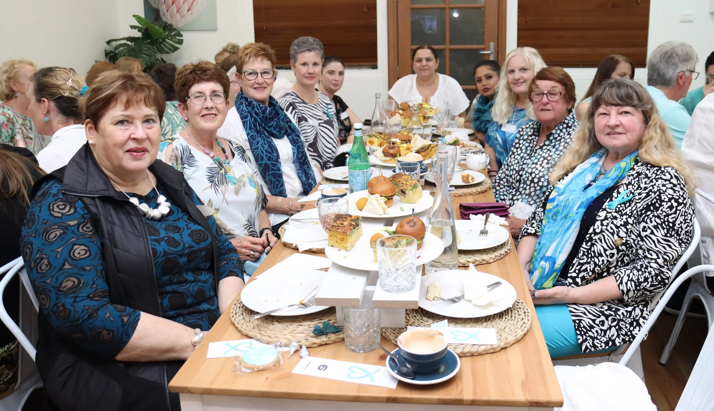 Group of women sitting around a dining table with food, drinks, and desserts, at a social gathering or celebration.