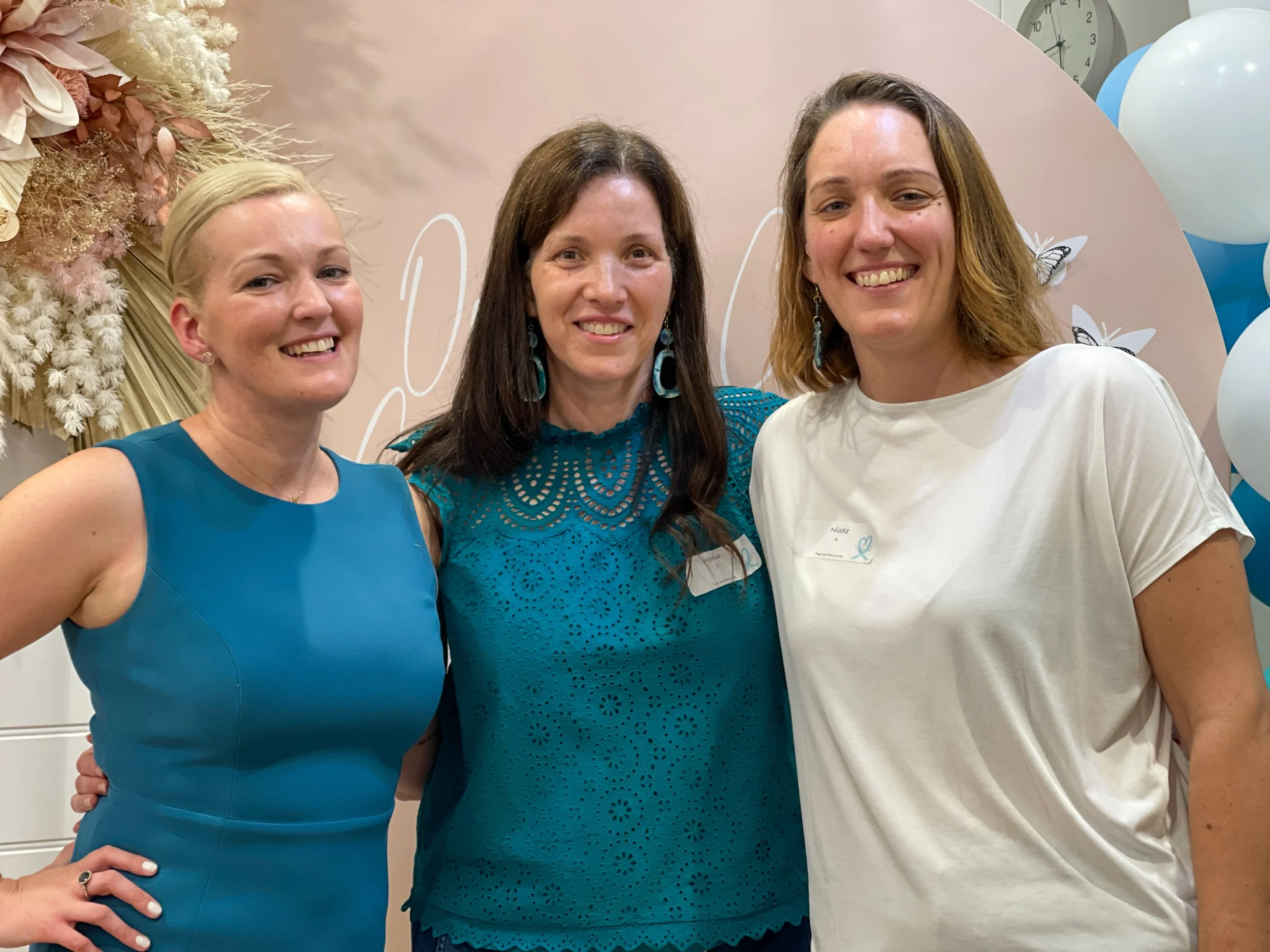 Three women smiling at the camera during a celebration with balloons and floral decorations in the background, one woman wearing a blue dress, another in a teal top, and the third in a white t-shirt.