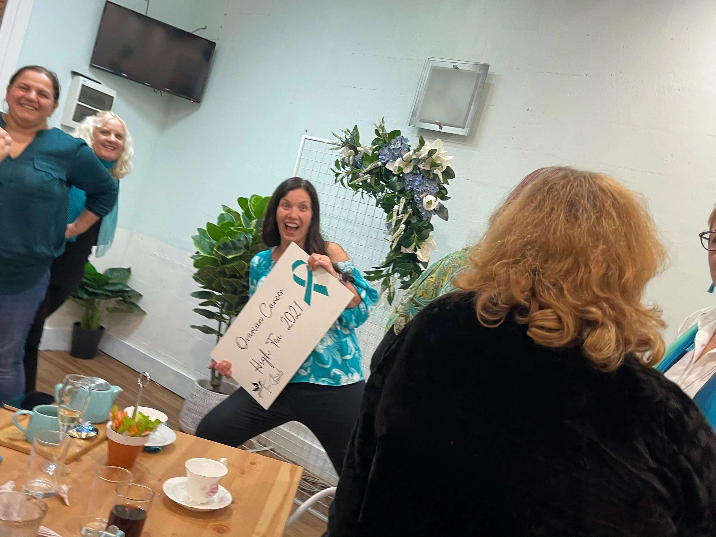 A group of women celebrating at an event, with one woman in the center holding a large check that says 'Ovarian Cancer Hope Tour 2021' and a teal ribbon symbolizing ovarian cancer awareness. The setting appears to be a casual gathering with a table o