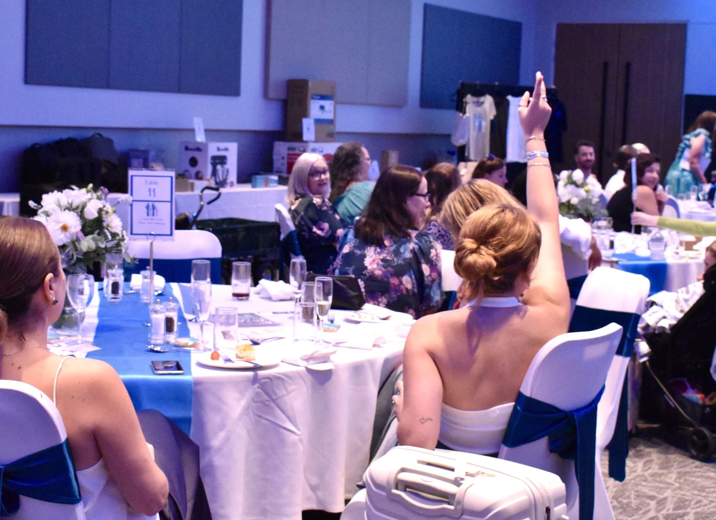 A woman with her hair up, wearing a strapless white dress, raising her hand in a recognition gesture during a formal gathering or event at a banquet hall.