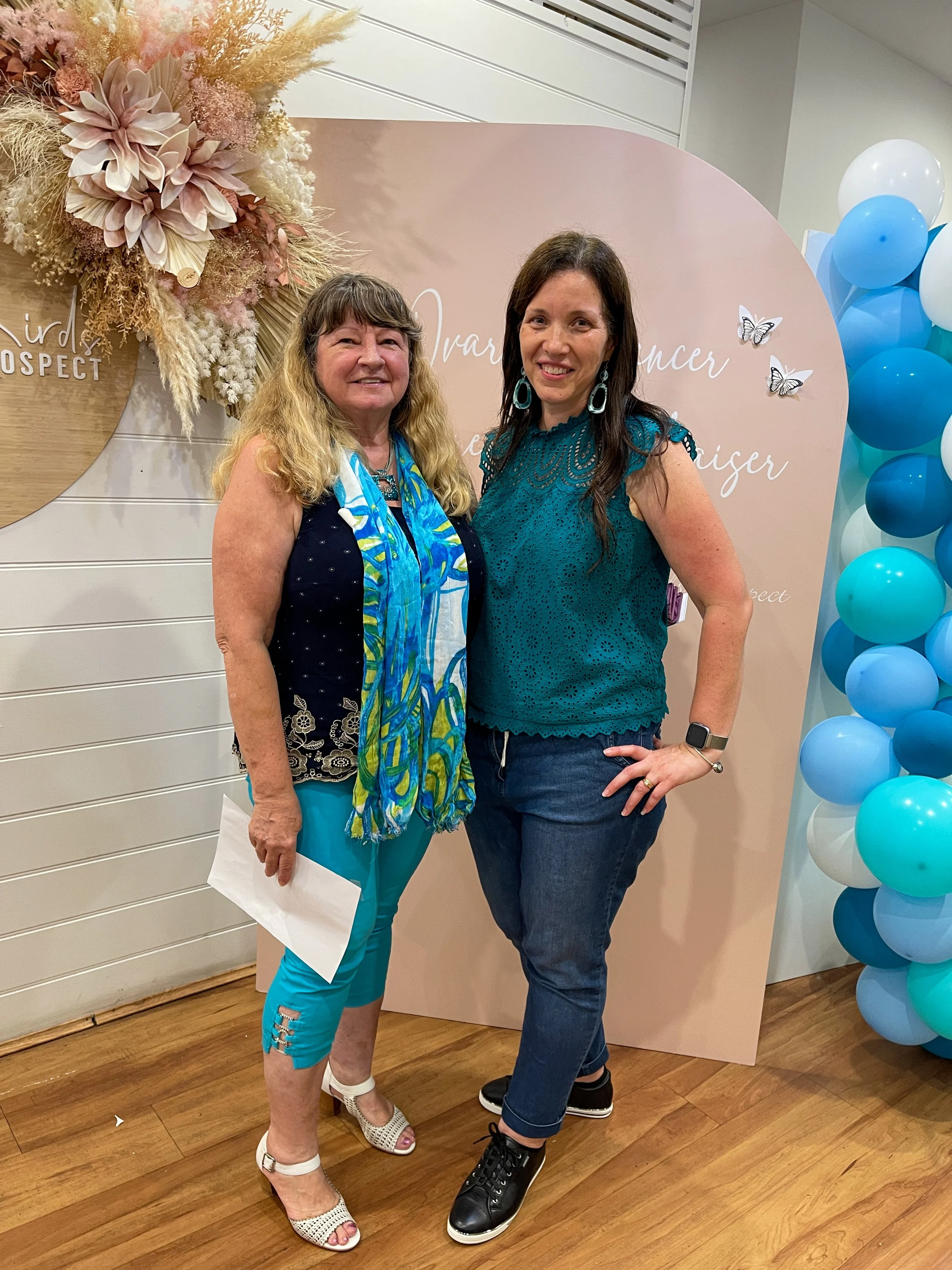 Two women standing together in front of a pink backdrop with butterflies and floral decorations, at an event celebrating women entrepreneurs, with blue and white balloons on the side.