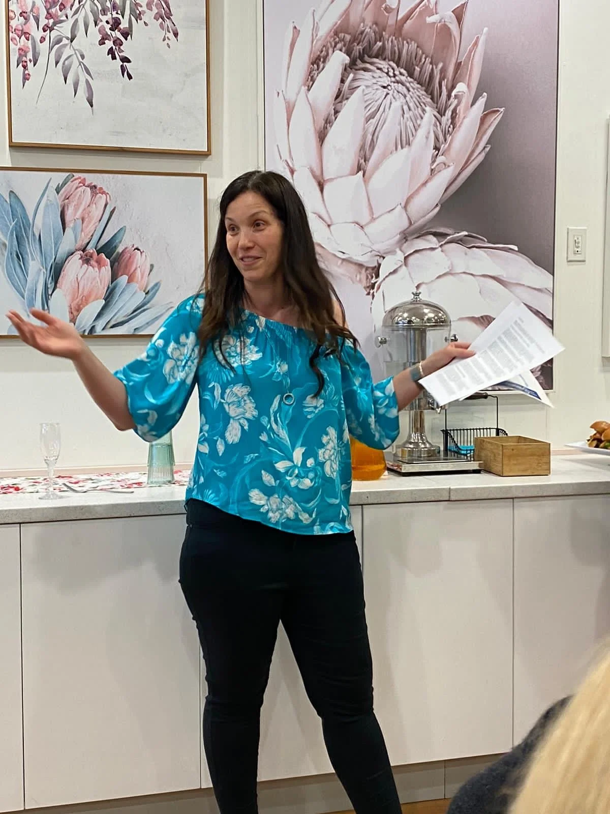 Woman giving a presentation at a social gathering, standing in front of floral artwork and a refreshment station, holding papers.