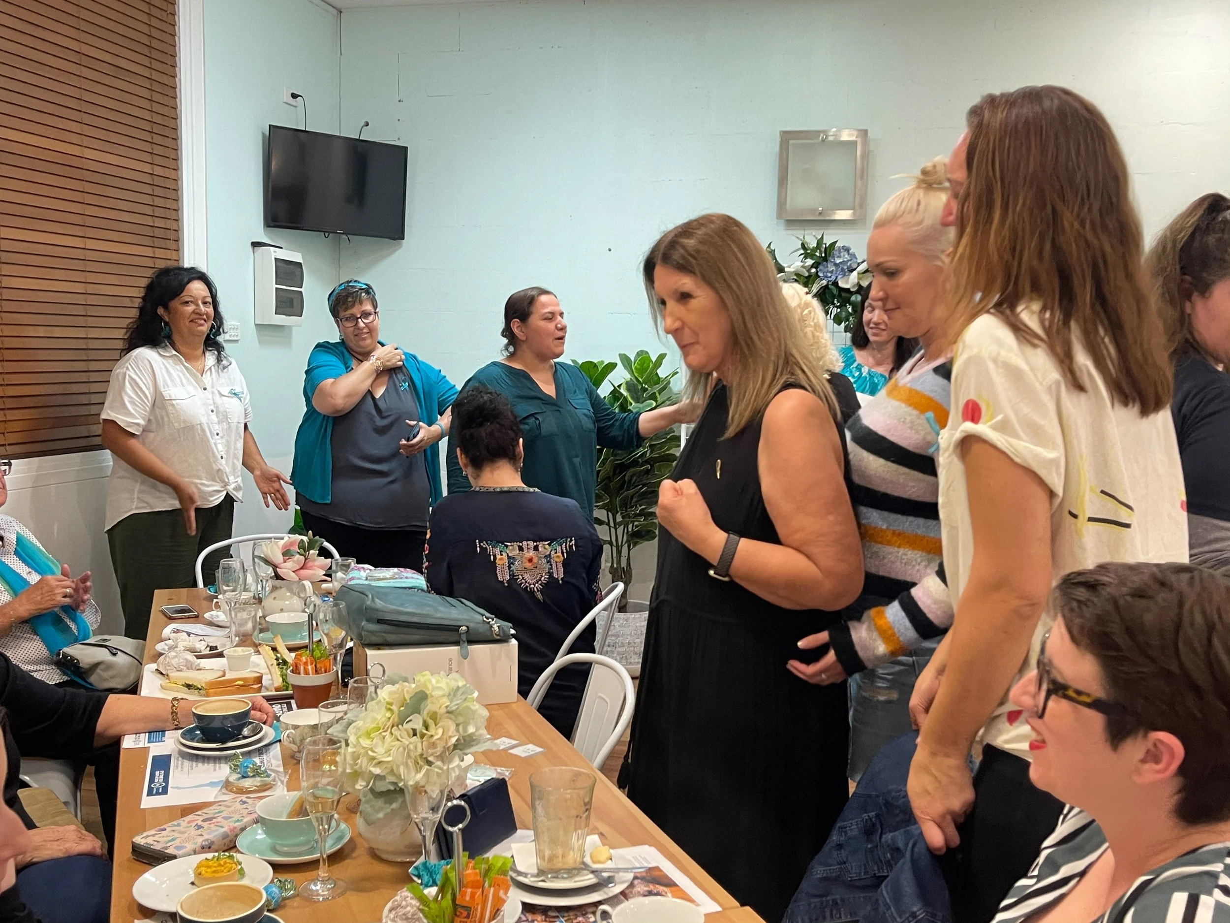 A group of women gathered in a meeting room, some standing and some seated at a table with food and drinks, participating in a social or professional gathering.