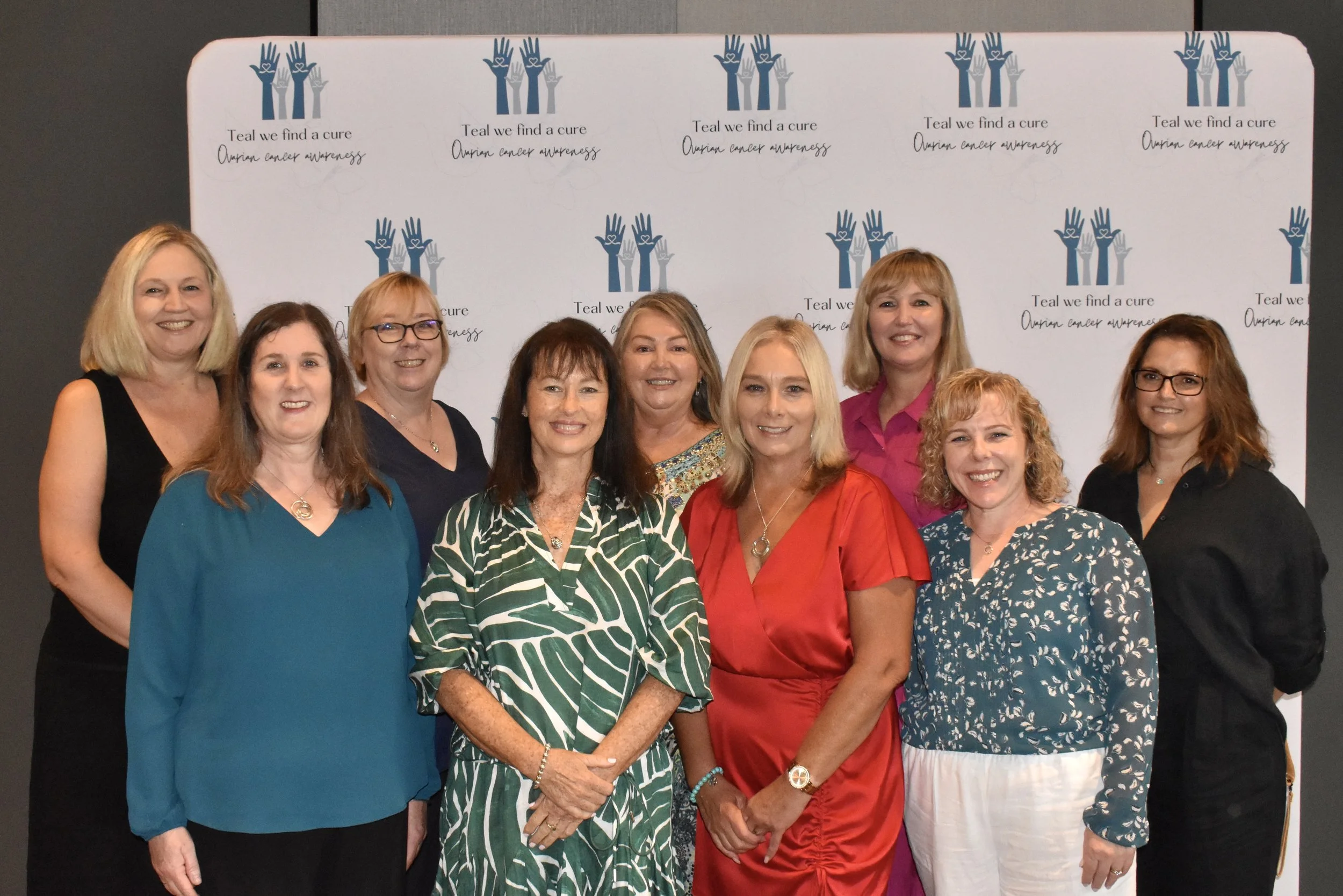 Group of ten women standing in front of a pink backdrop with the text 'Teal we find a cure ovarian cancer awareness' and a logo with hands. They are dressed in colorful casual and semi-formal clothing, smiling at the camera.