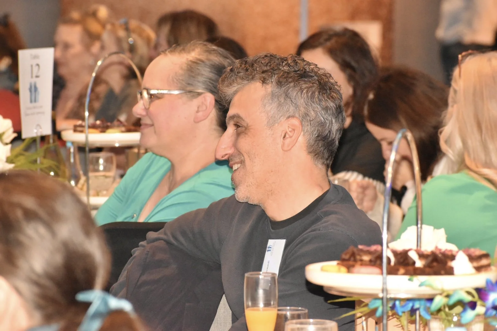 People sitting at a table during a gathering or party, smiling, with a tiered tray of desserts and glasses of drinks in front of them.