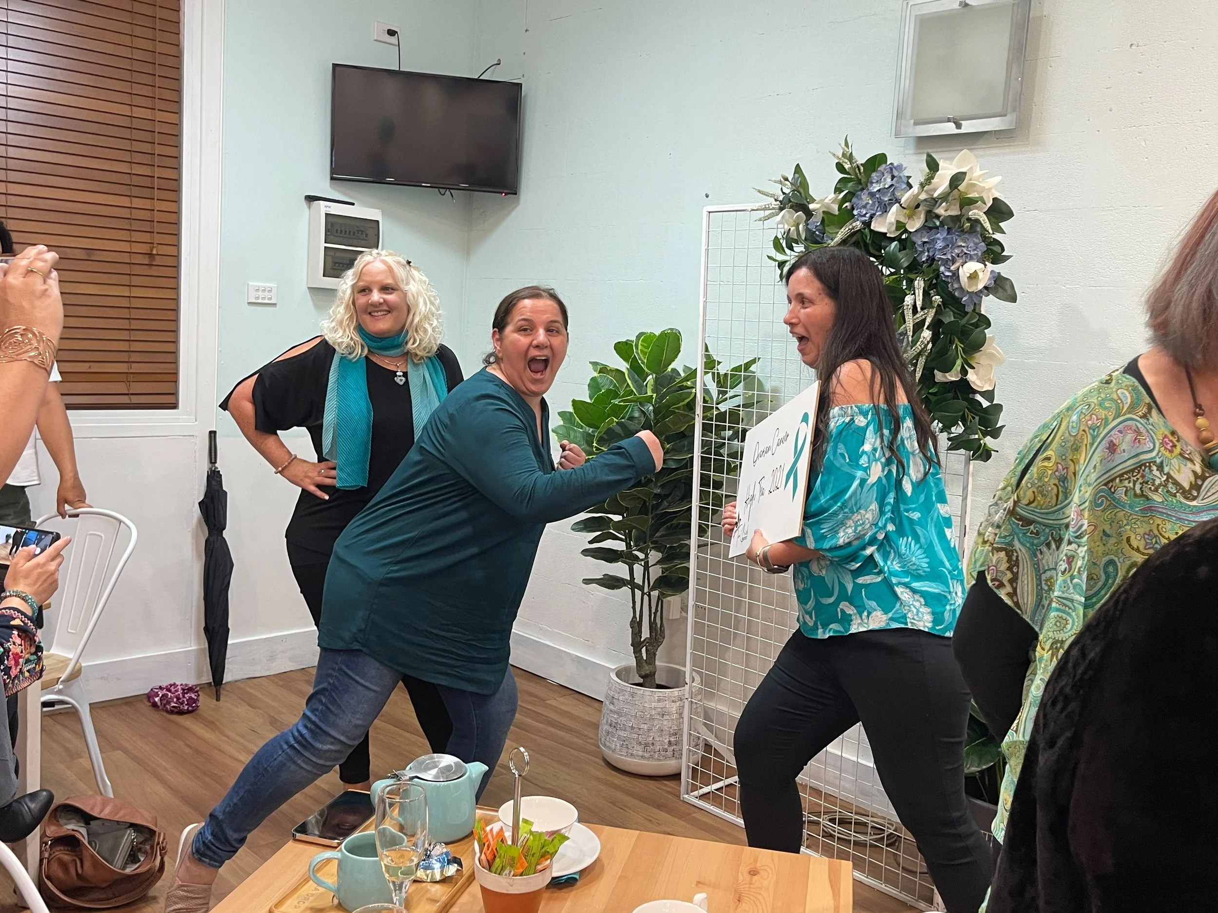 Women celebrating at a gathering, one woman appears to be receiving an award or recognition while others cheer and smile, with a whiteboard and large floral arrangement in the background.
