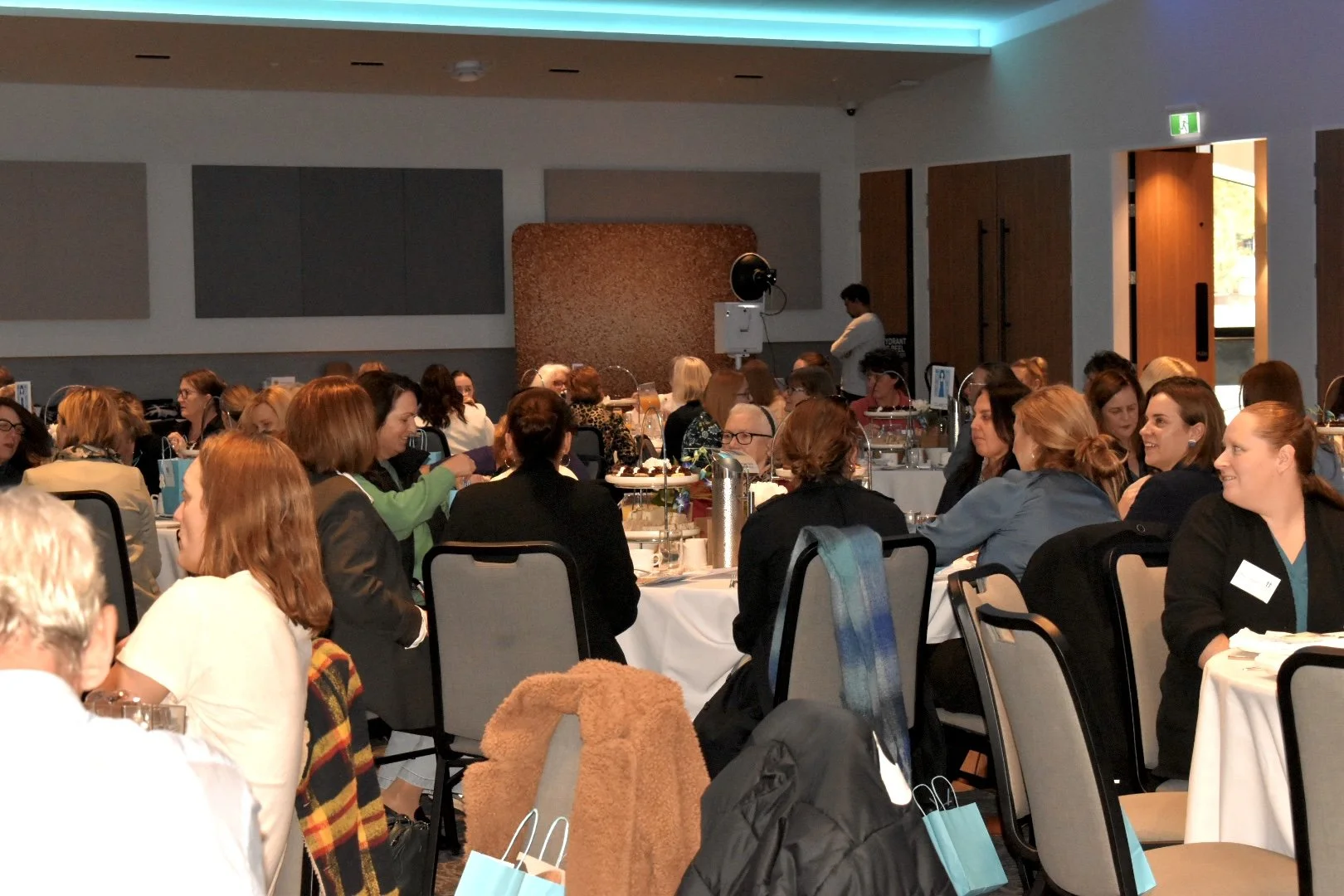 A large group of women and a few men seated at round tables during a conference or event inside a modern, well-lit room.