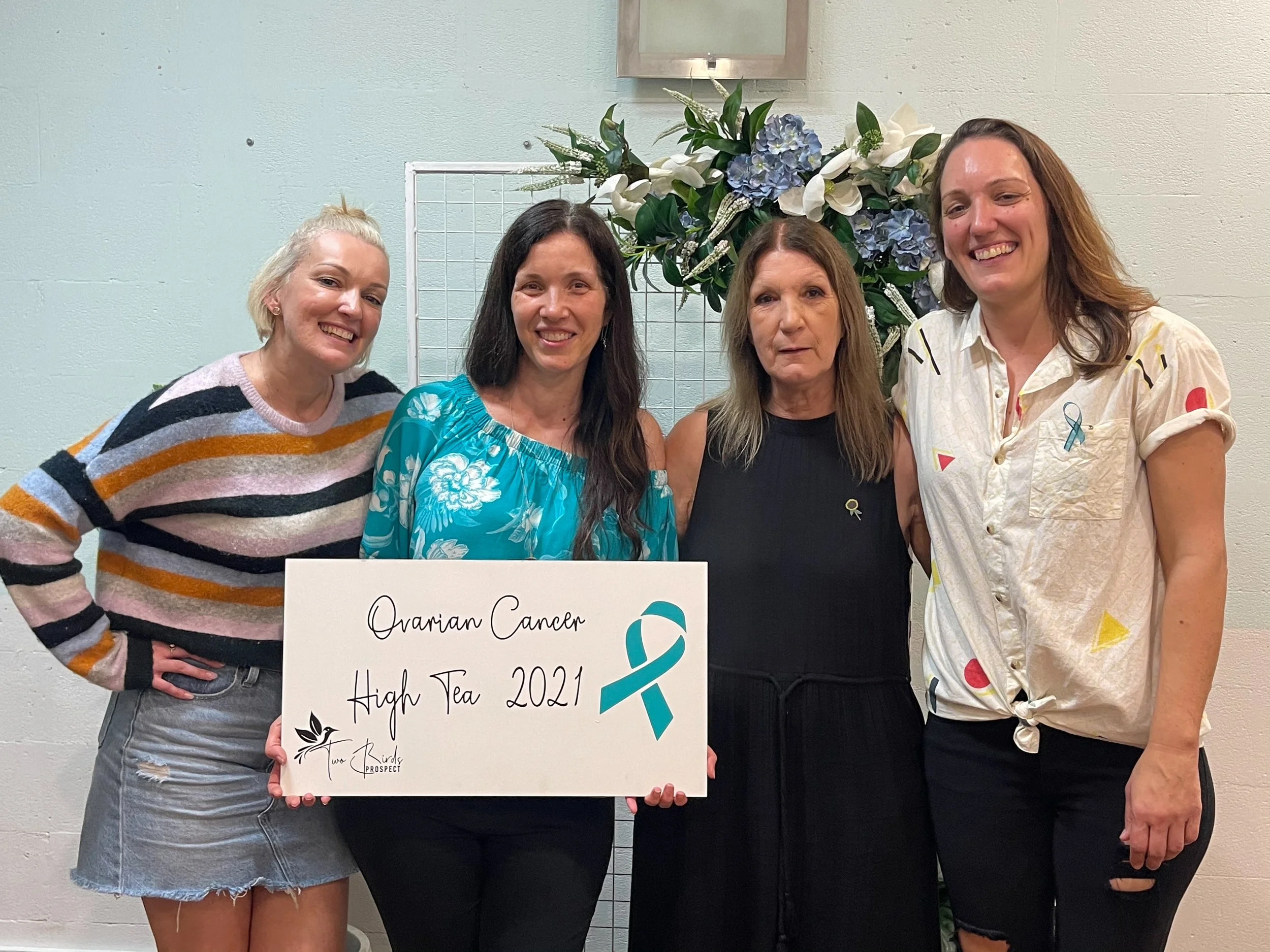 Four women standing together indoors, holding a white sign with black and blue lettering that reads "Ovarian Cancer High Tea 2021," with a blue awareness ribbon and a small black bird logo, and a large floral arrangement with white and blue flowers b