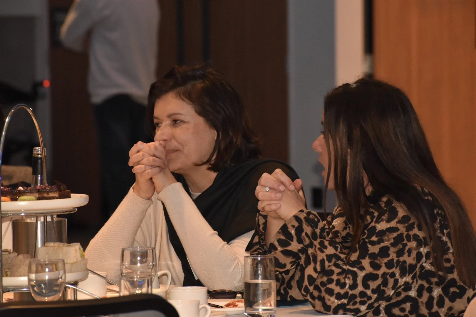 Two women sitting at a table, one with hands clasped near her face, engaging in conversation, with food and drinks on the table.