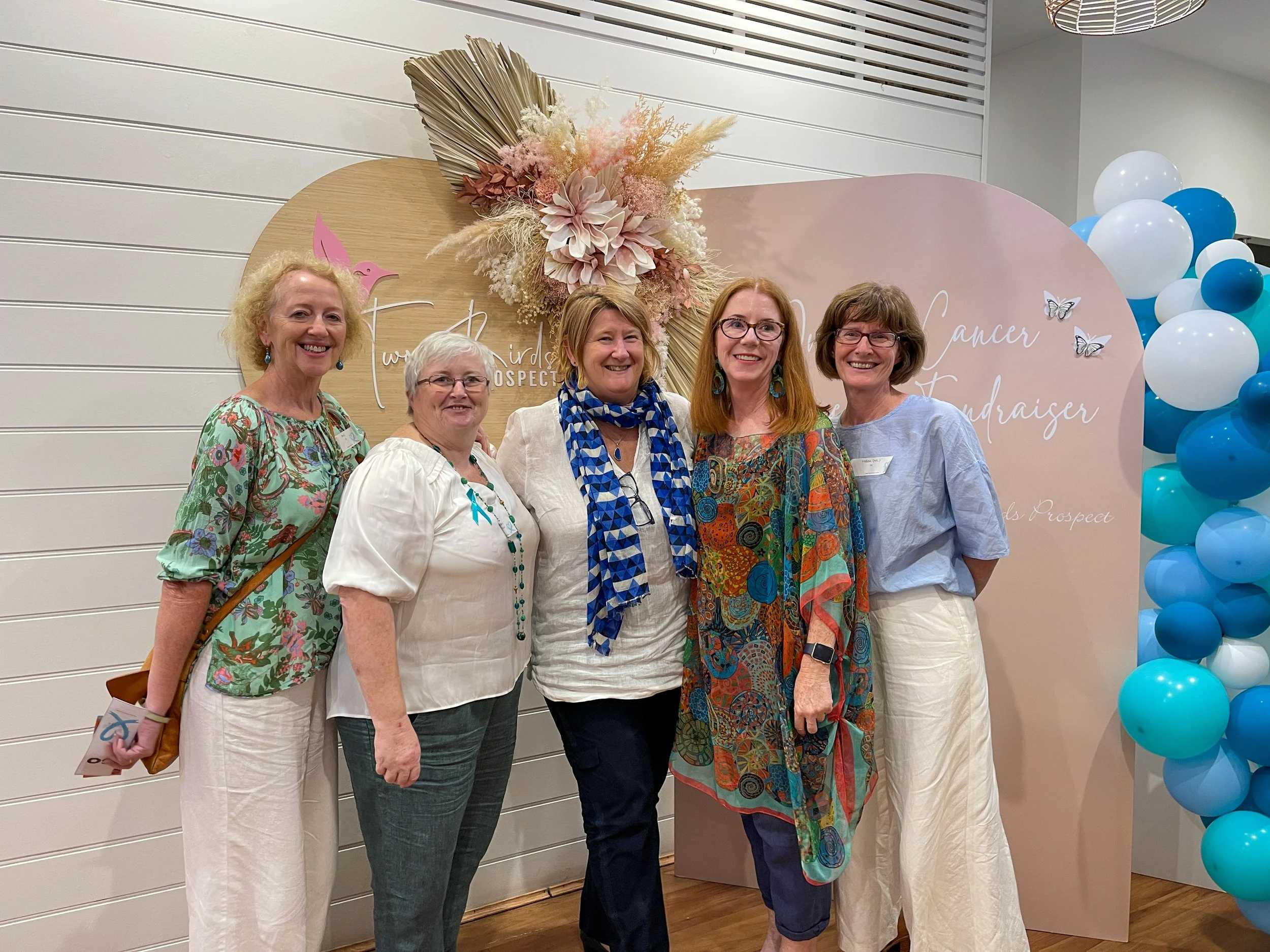 Five women standing together at a breast cancer fundraiser event, smiling, with a pink backdrop decorated with flowers and balloons.