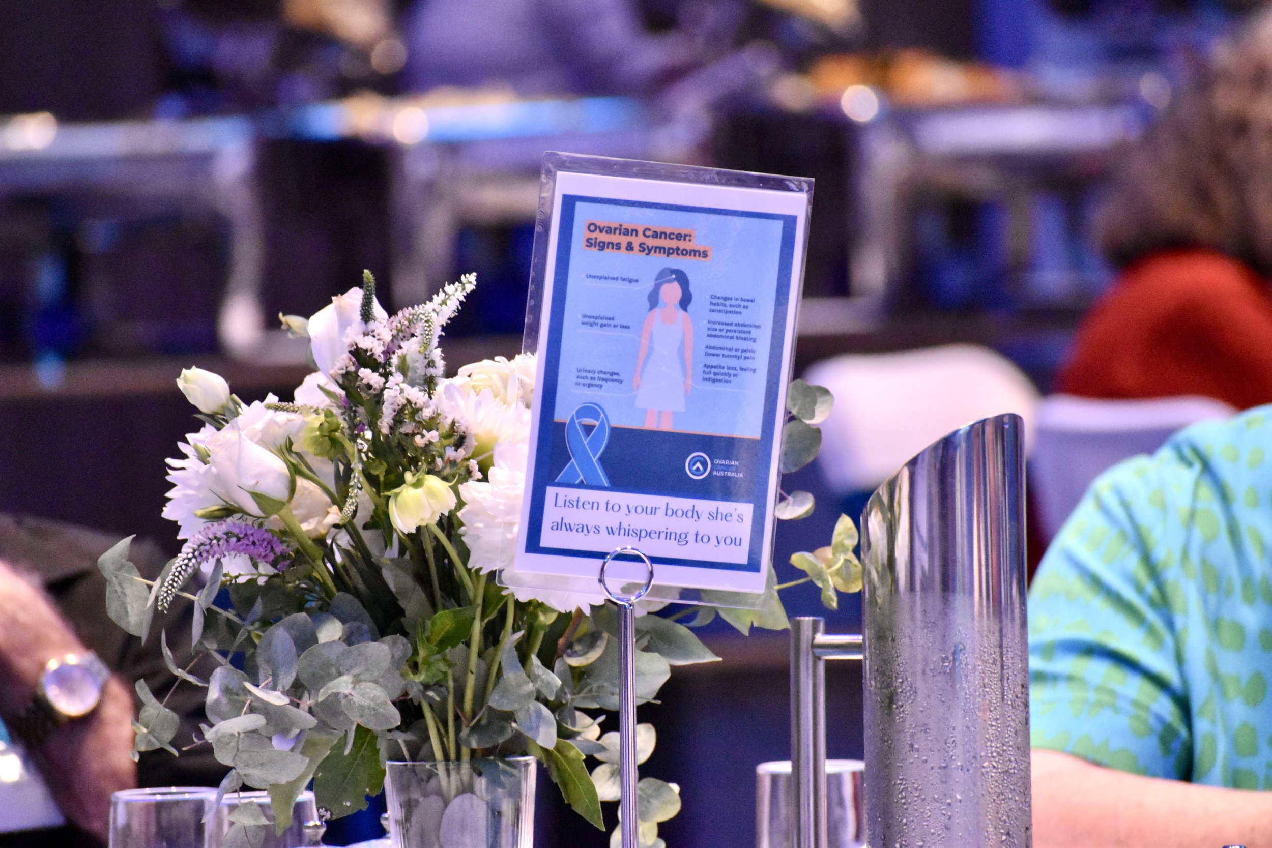 A table at an event features a bouquet of white flowers and an informational sign about ovarian cancer symptoms. The event appears to be indoors, with several chairs and people in the background.
