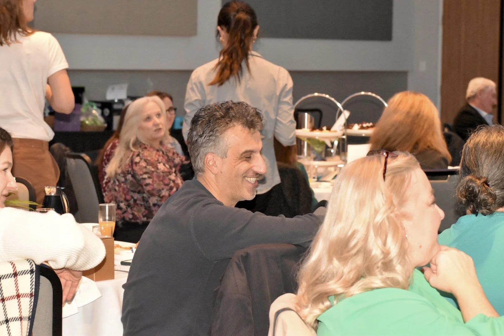 Group of people sitting at tables in a conference room or banquet hall, engaged in conversation and smiling, with food and drinks on the tables.