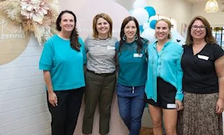 Five women standing together indoors, smiling for a photo at a celebration event with balloons in the background.
