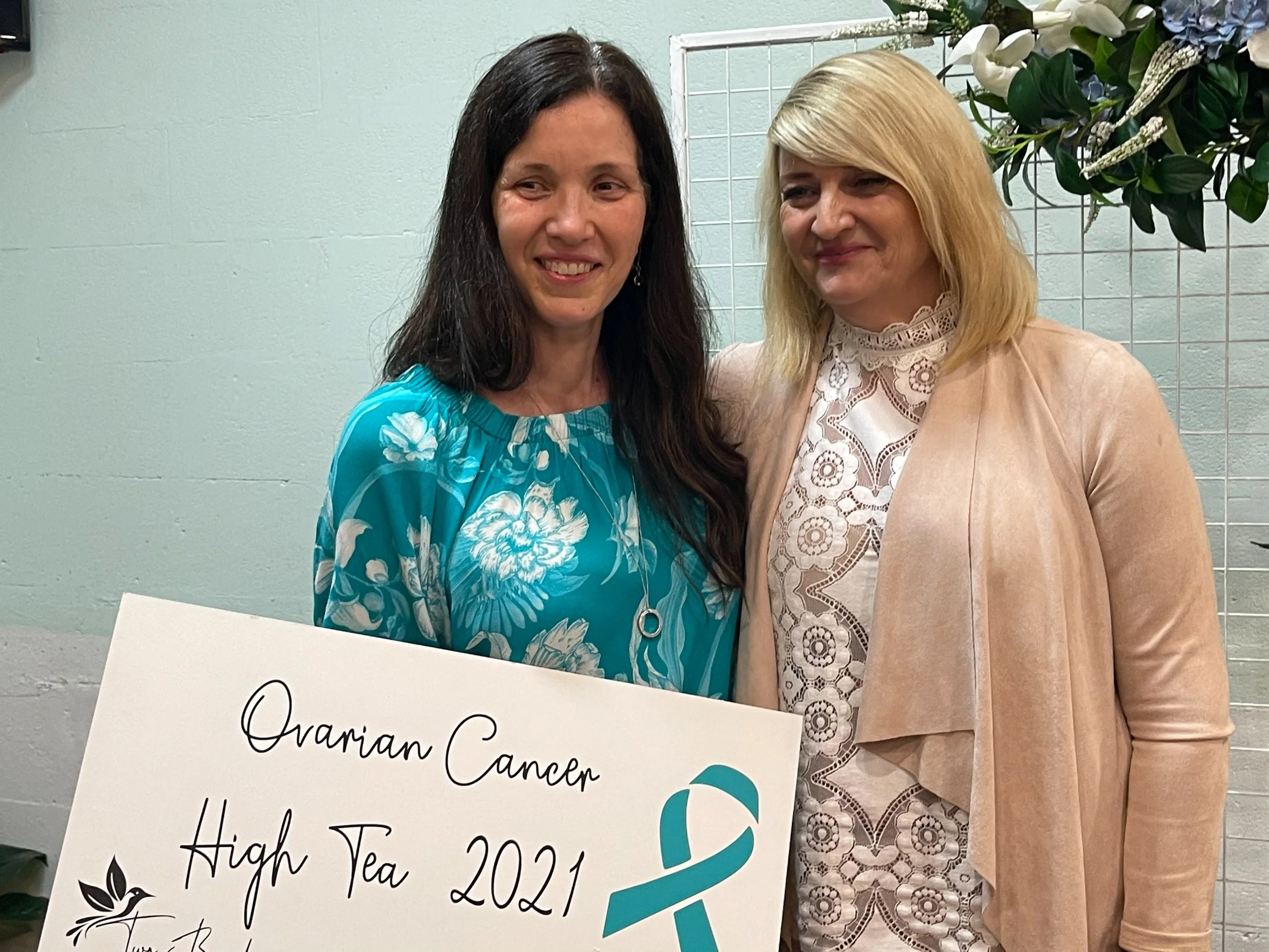 Two women standing close together, smiling, at an ovarian cancer awareness event in 2021. One woman is holding a sign that reads "Ovarian Cancer High Tea 2021" with a teal ribbon symbol and a floral design.