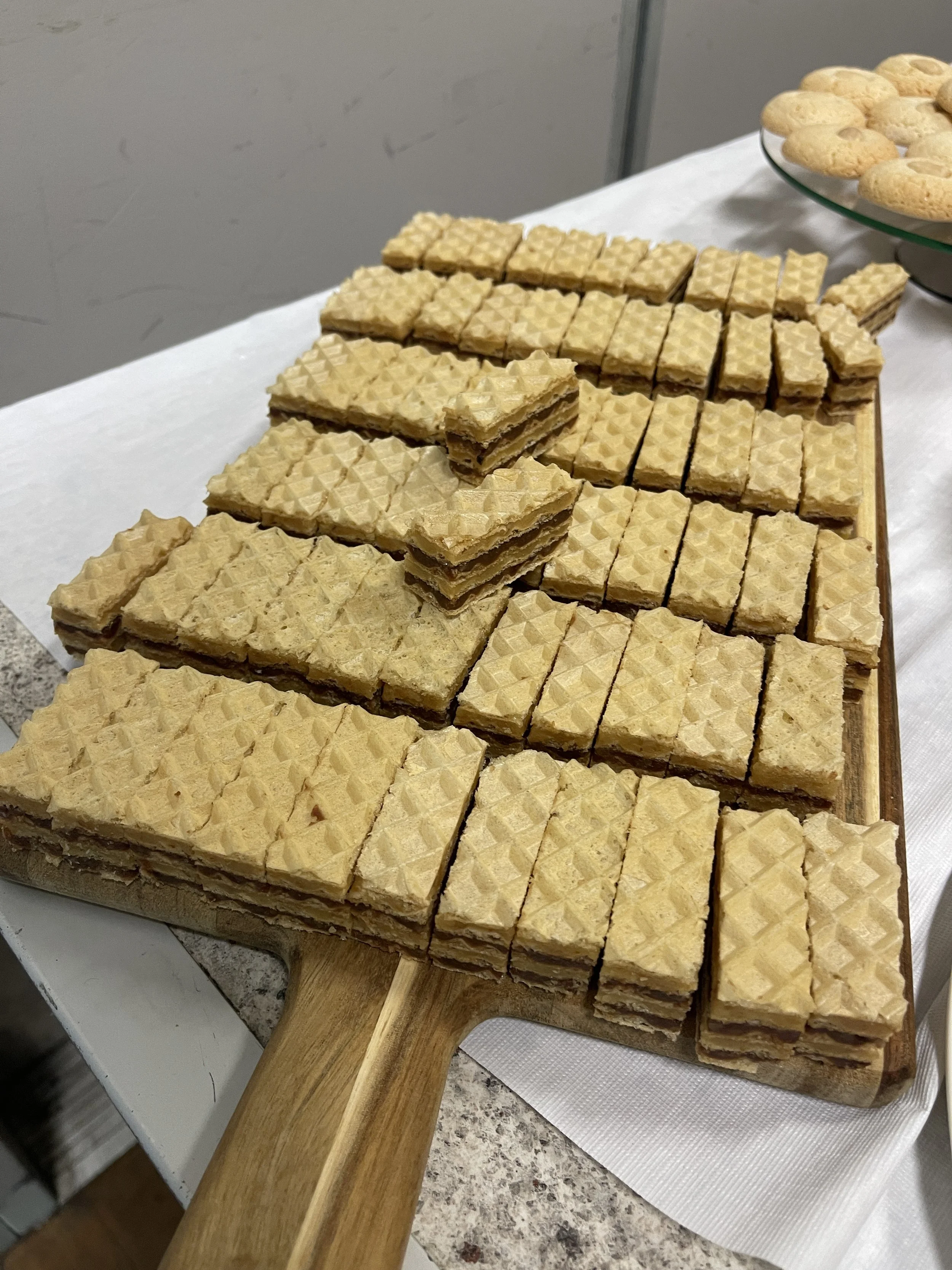 A wooden paddle-shaped tray with layered wafer cookies and some pieces stacked, set on a white cloth surface.