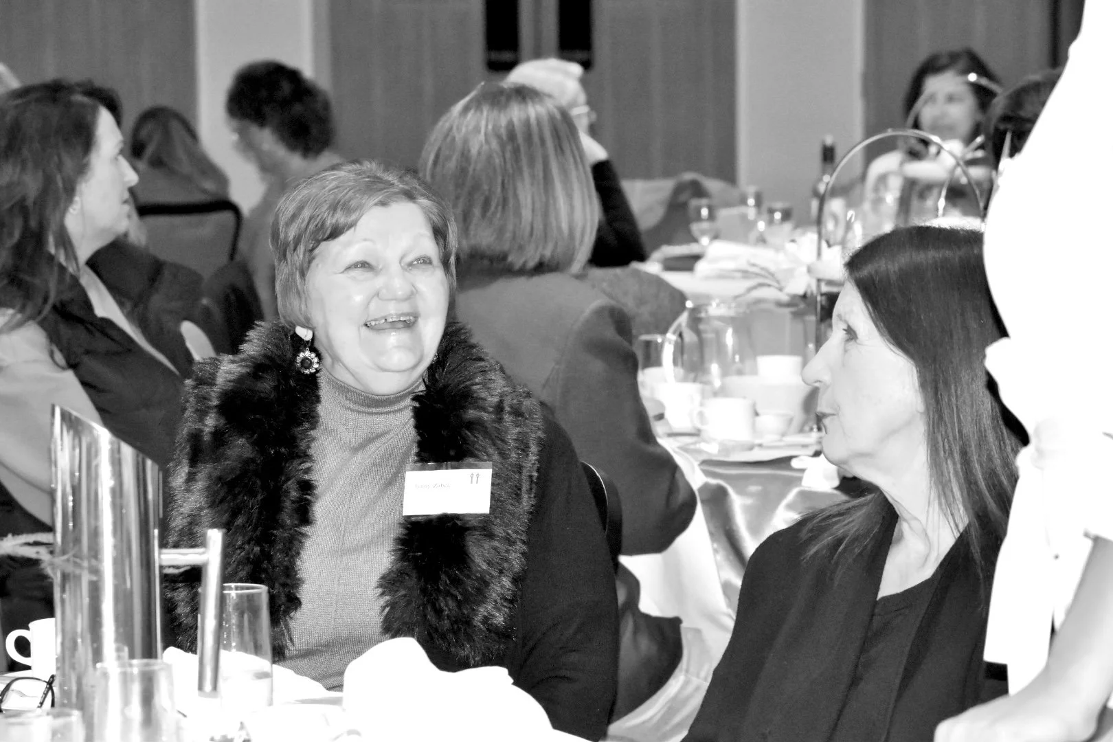 Women at a social gathering or conference, seated at round tables with tableware, engaged in conversation.