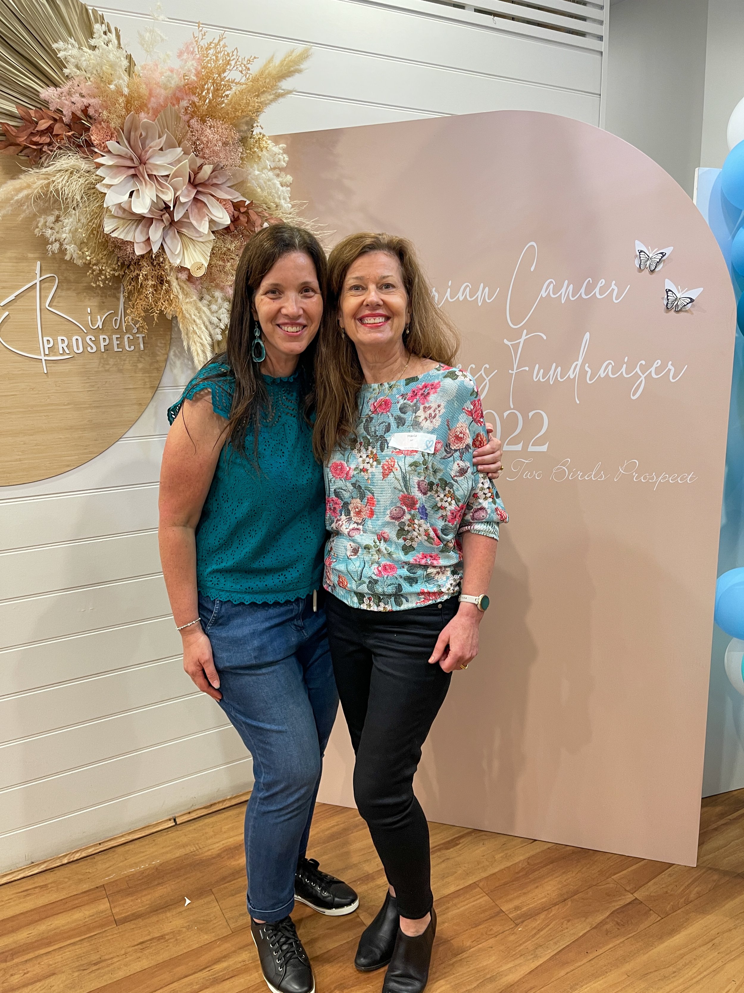 Two women smiling and posing together at a breast cancer awareness fundraiser event with a pink backdrop and floral decoration.