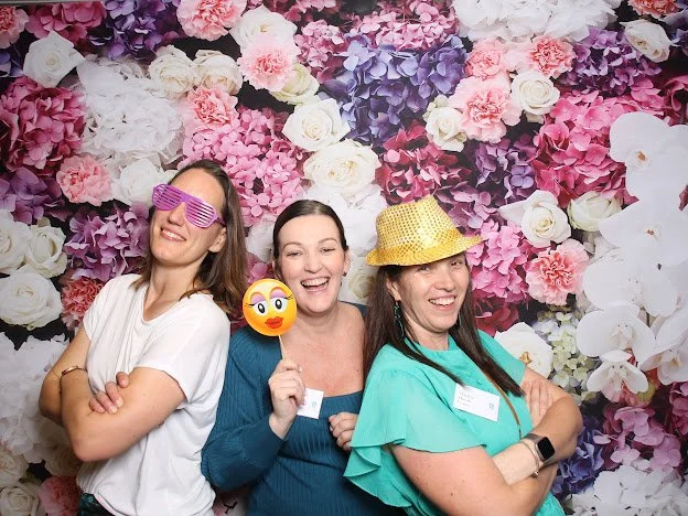 Three women smiling and posing for a photo at a party with a floral backdrop of pink, purple, white, and peach flowers. They are wearing fun accessories, including large shutter shades, a yellow hat, and holding an emoji sign.