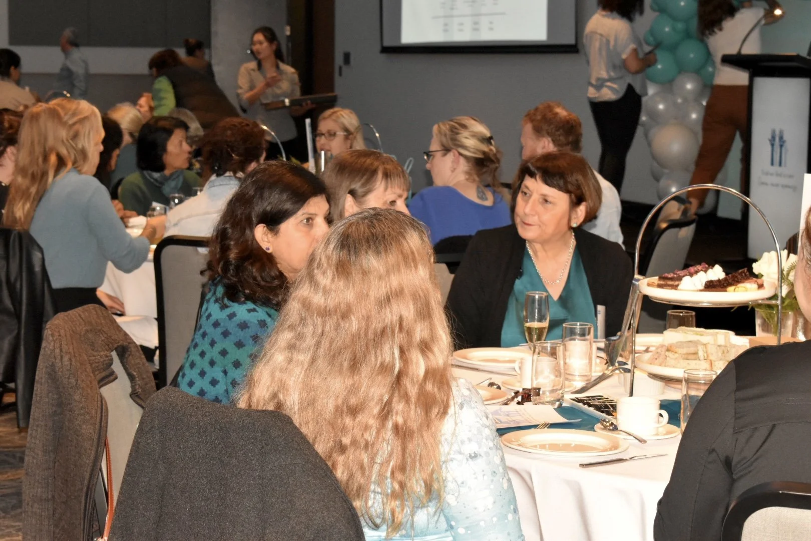 Women sitting around a dinner table at a formal event, engaged in conversations, with food and drinks on the table, and a stage with a presentation screen and balloon decorations in the background.