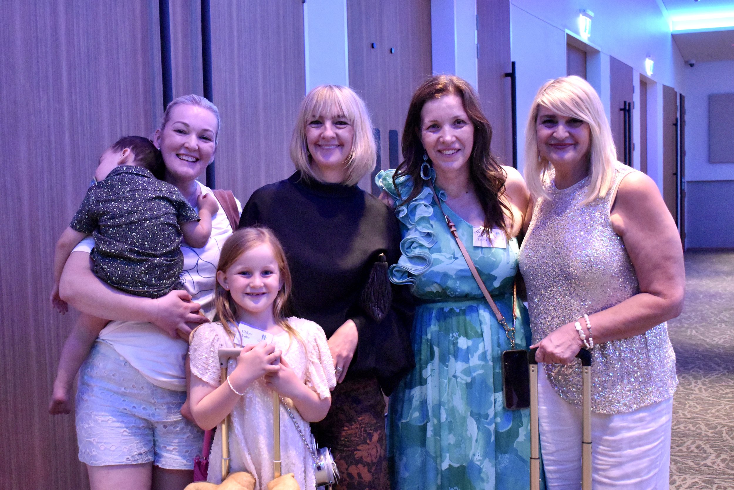 Group of five women and two young girls smiling at a social event in a well-lit room with wooden walls.