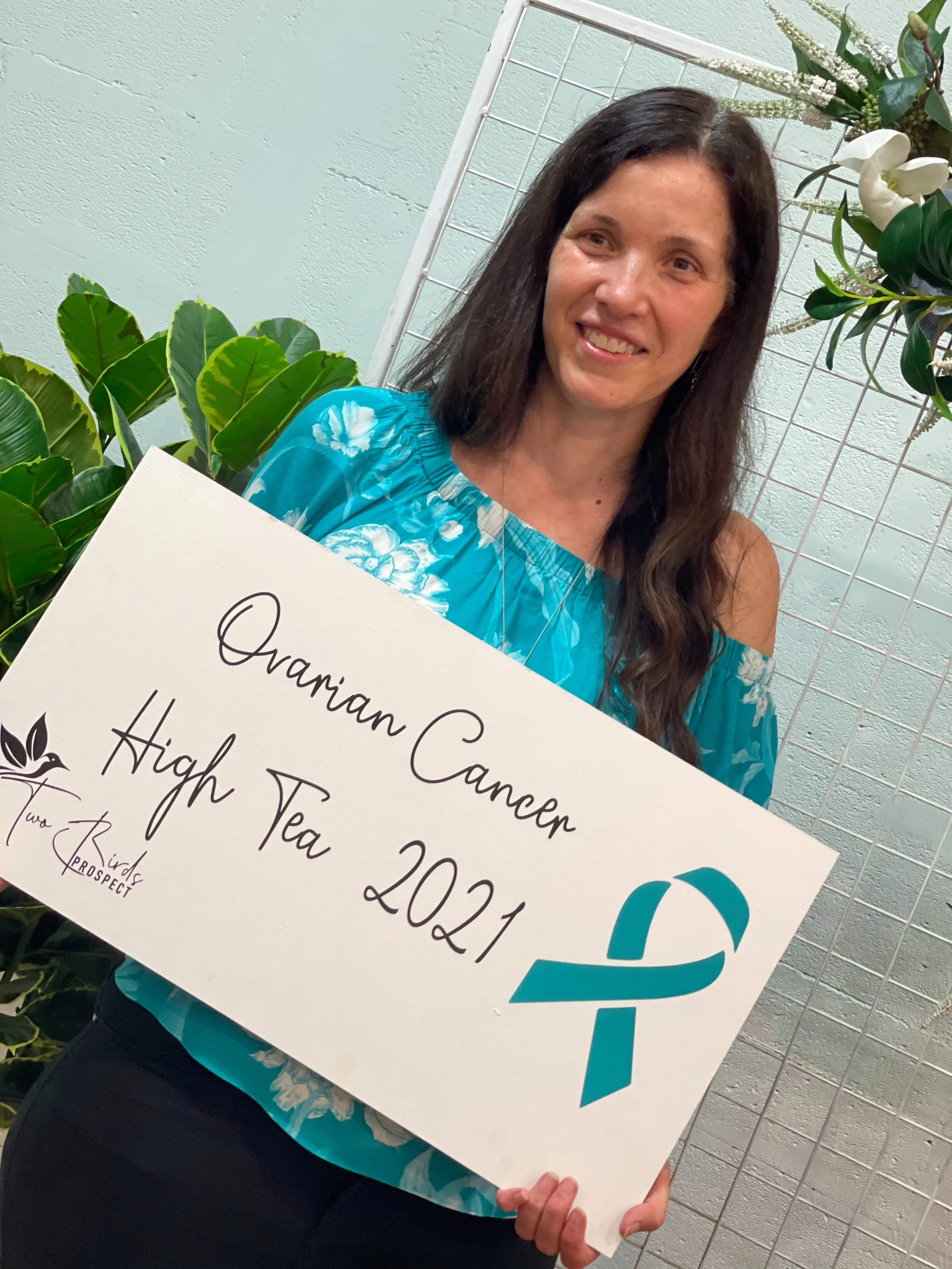 A woman with dark hair and a turquoise floral blouse holding a sign that reads 'Ovarian Cancer High Tea 2021' with a teal ribbon, standing in front of a white grid and green plants.