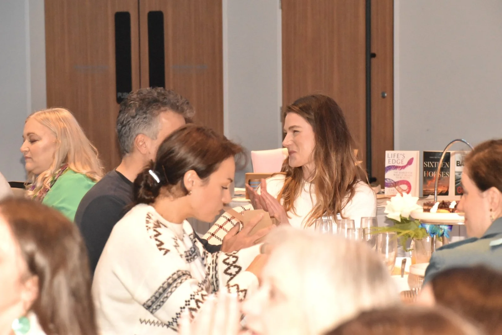 People sitting at a banquet table, engaging in conversation, with books and floral arrangement in the background.