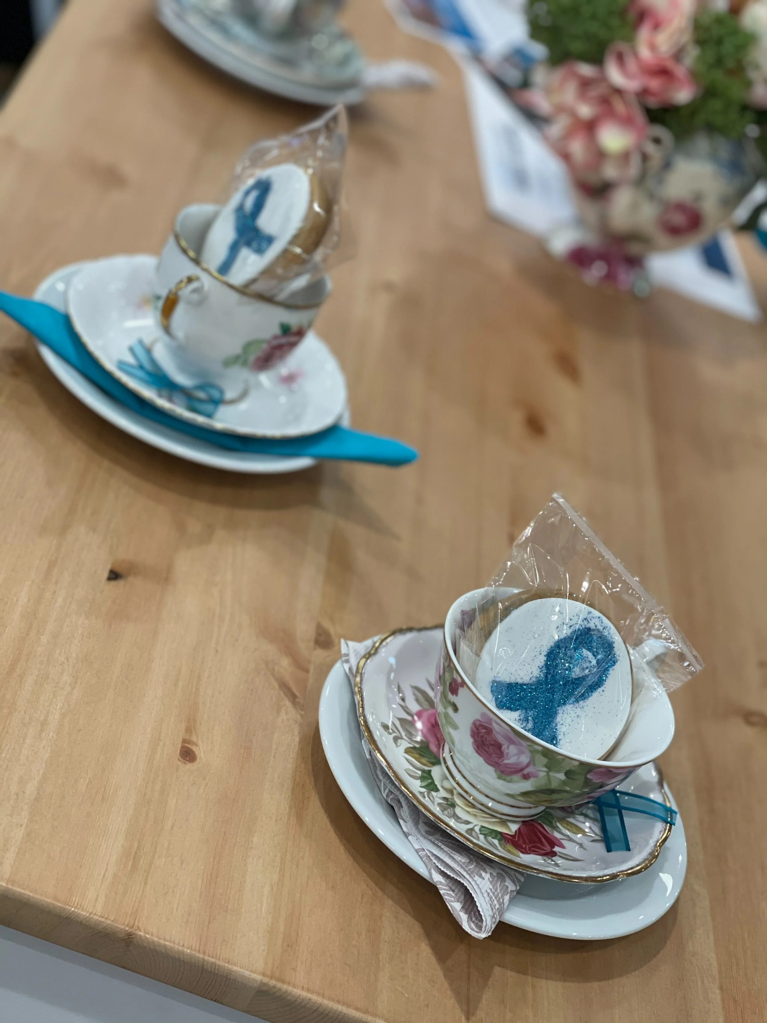 Two tea cups with saucers, each with a blue sparkly treat wrapped in clear plastic, placed on napkins and small plates on a wooden table, with a vase of pink flowers in the background.