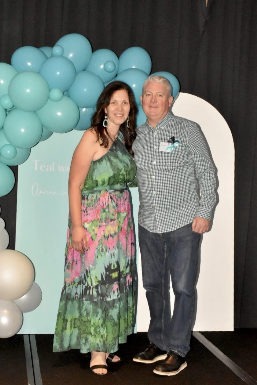 A woman in a colorful green and pink dress and a man in a checkered shirt and jeans standing together at an event with a backdrop of teal balloons and a white and teal sign.