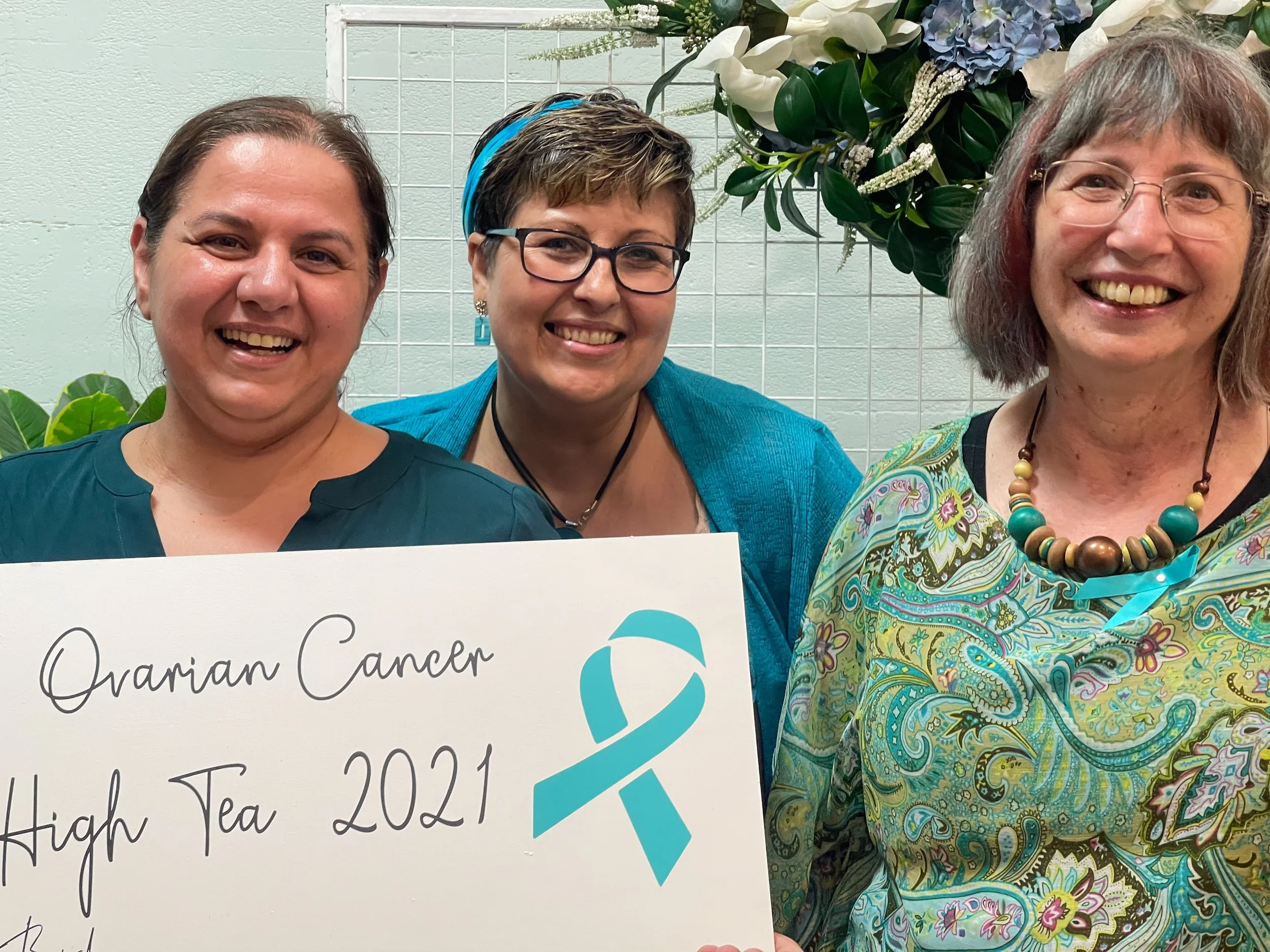 Three women smiling and standing together, holding a sign that reads 'Ovarian Cancer High Tea 2021' with a teal ribbon. They are indoors, with greenery and flowers in the background.