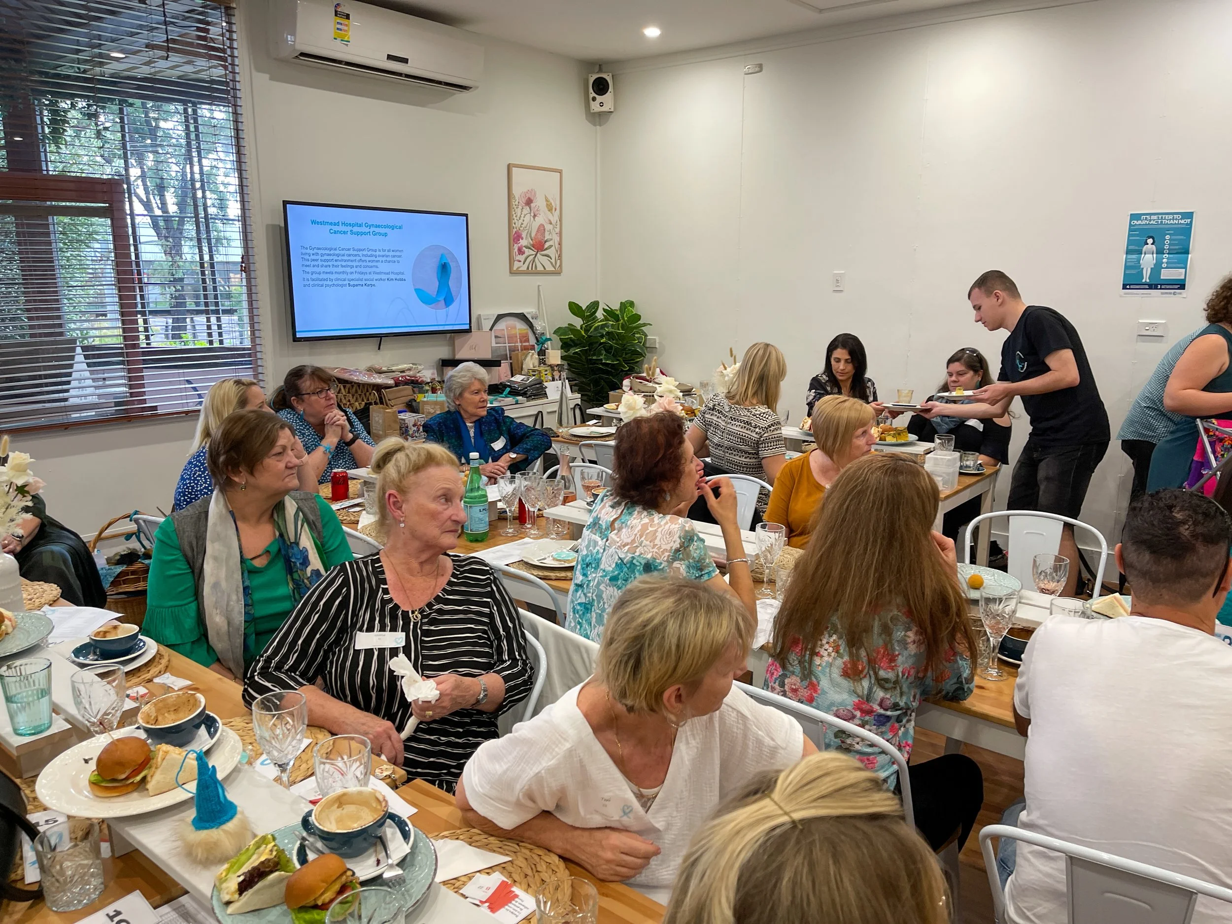 A group of people seated at tables in a room, attending a meeting or gathering, with a speaker serving food. There is a screen on the wall displaying a presentation related to a hospital gynecological cancer support group, and several posters and pla