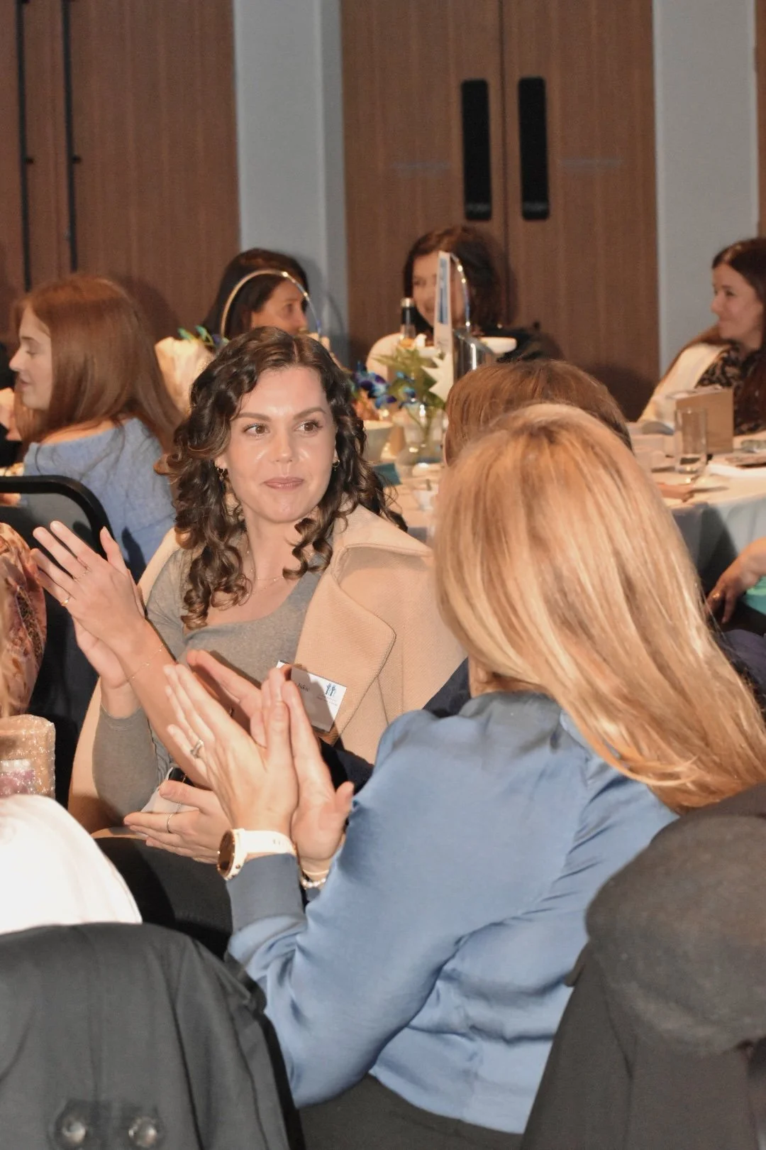 Women seated at tables, engaged in conversation during an event or conference, with some clapping and others listening.