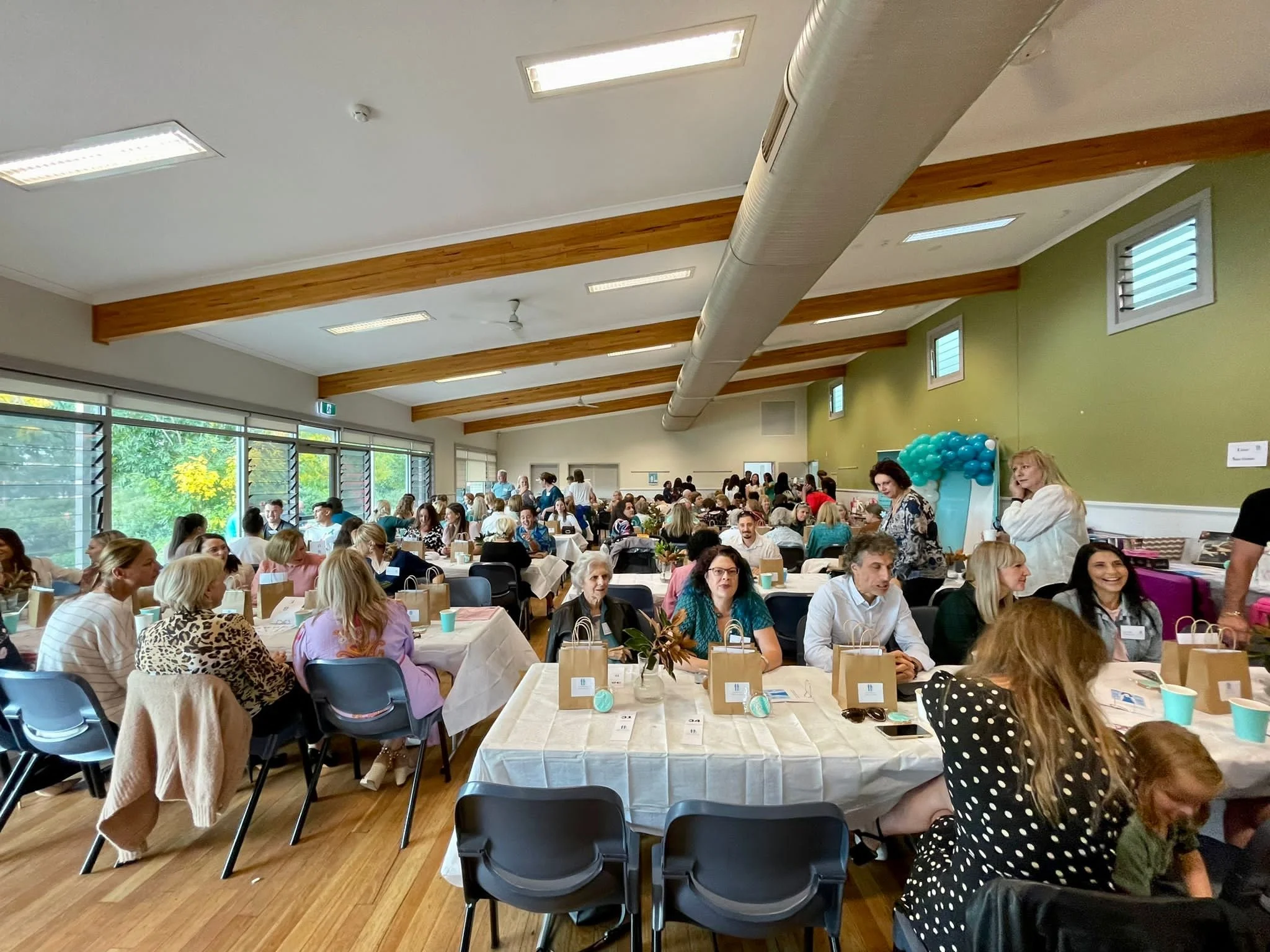 A large gathering of people seated at long tables in a spacious room with wooden beams on the ceiling and large windows letting in natural light. The event appears to be a social or professional gathering with decorations including balloons.