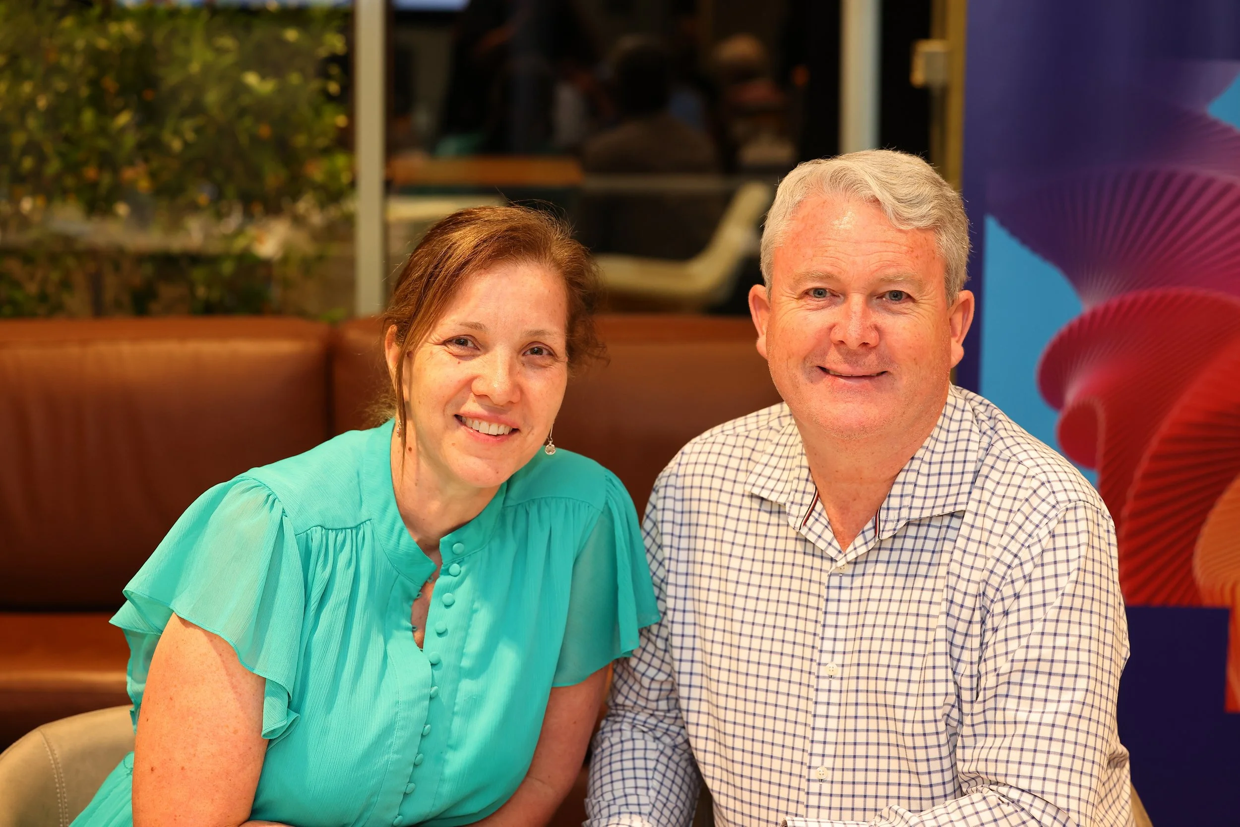 A smiling woman in a turquoise blouse and a smiling man in a checkered shirt sitting at a table in a restaurant or cafe with a colorful abstract mural and a window showing outdoor greenery in the background.