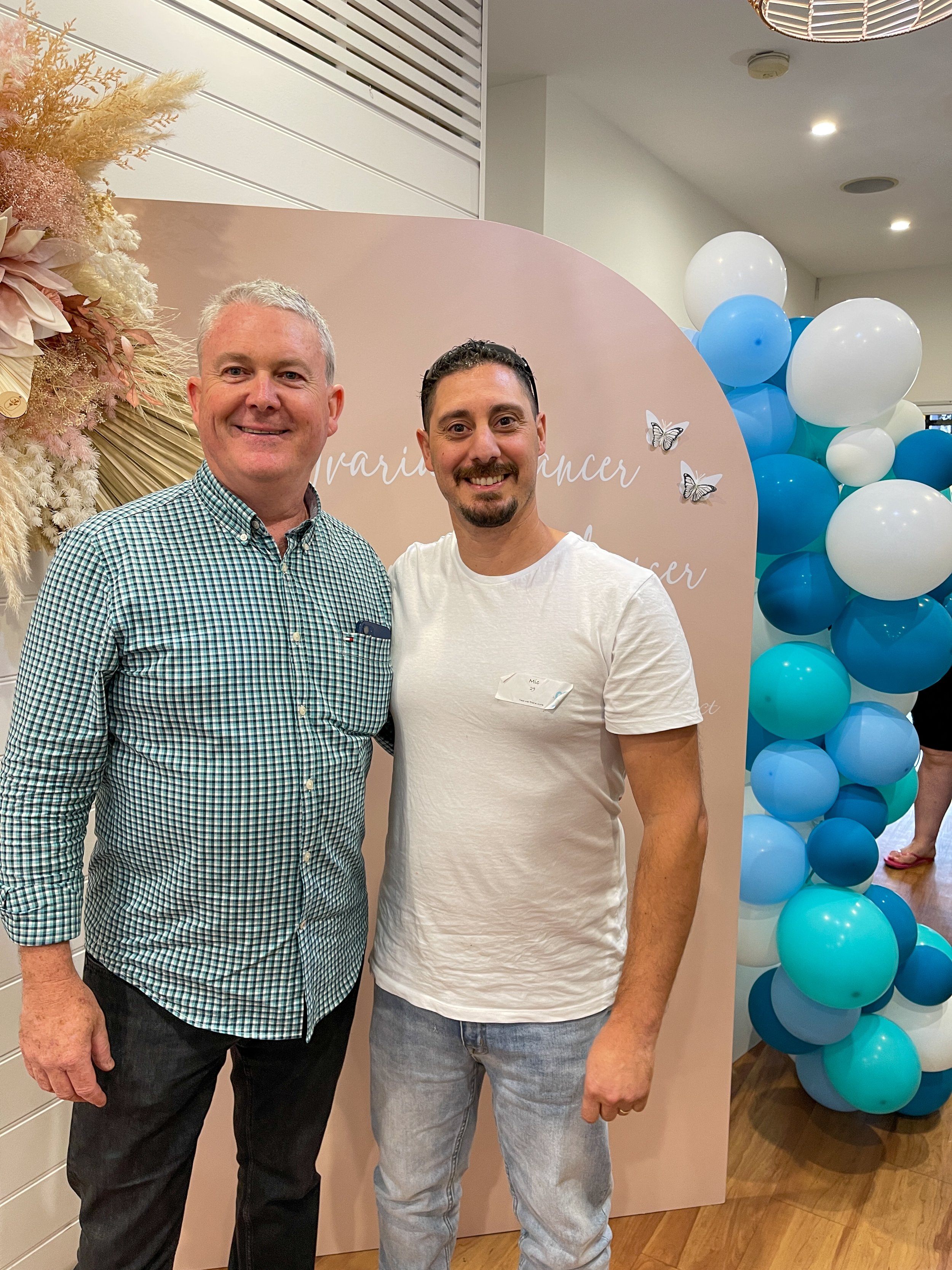 Two men smiling at an event with balloon decorations and a backdrop featuring butterflies and text.