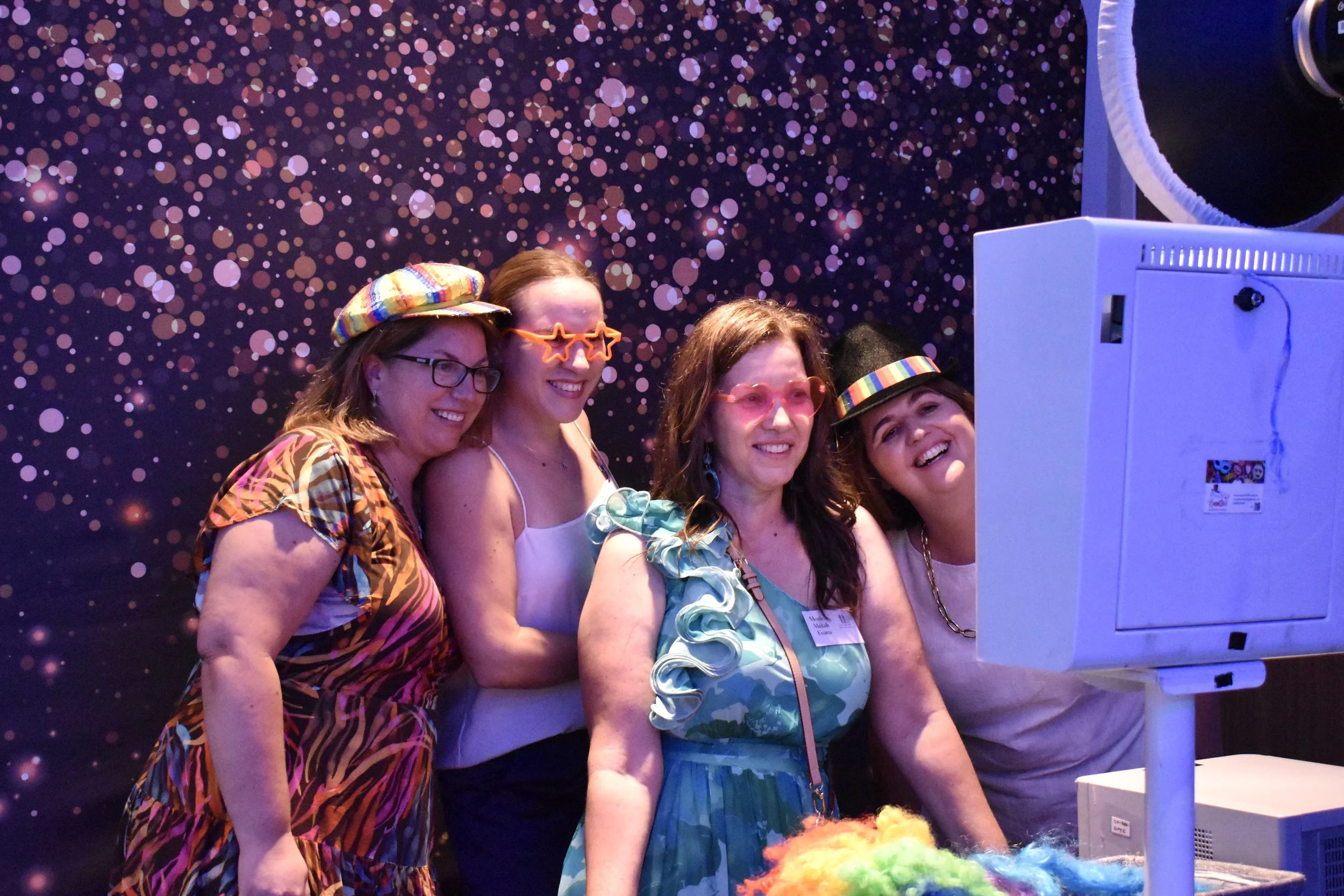 Four women dressed in colorful, festive clothing and accessories, wearing glasses and funky glasses, are gathered together looking at a large screen at an event with a starry background.