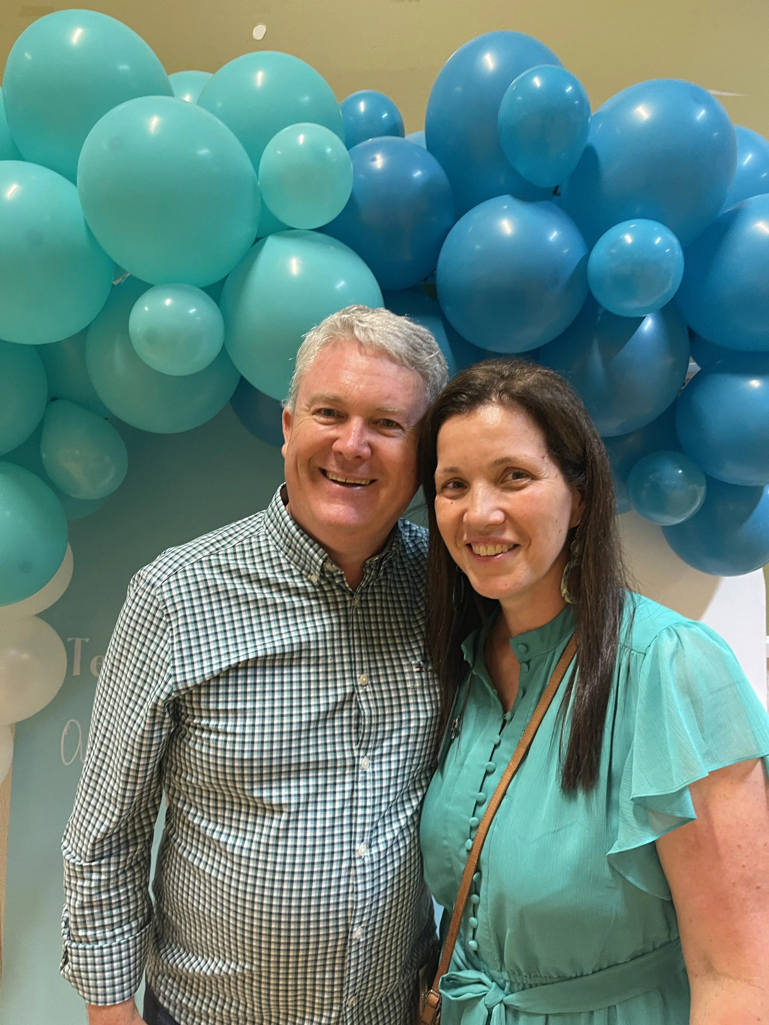 A smiling man and woman standing together in front of a backdrop of blue and teal balloons.