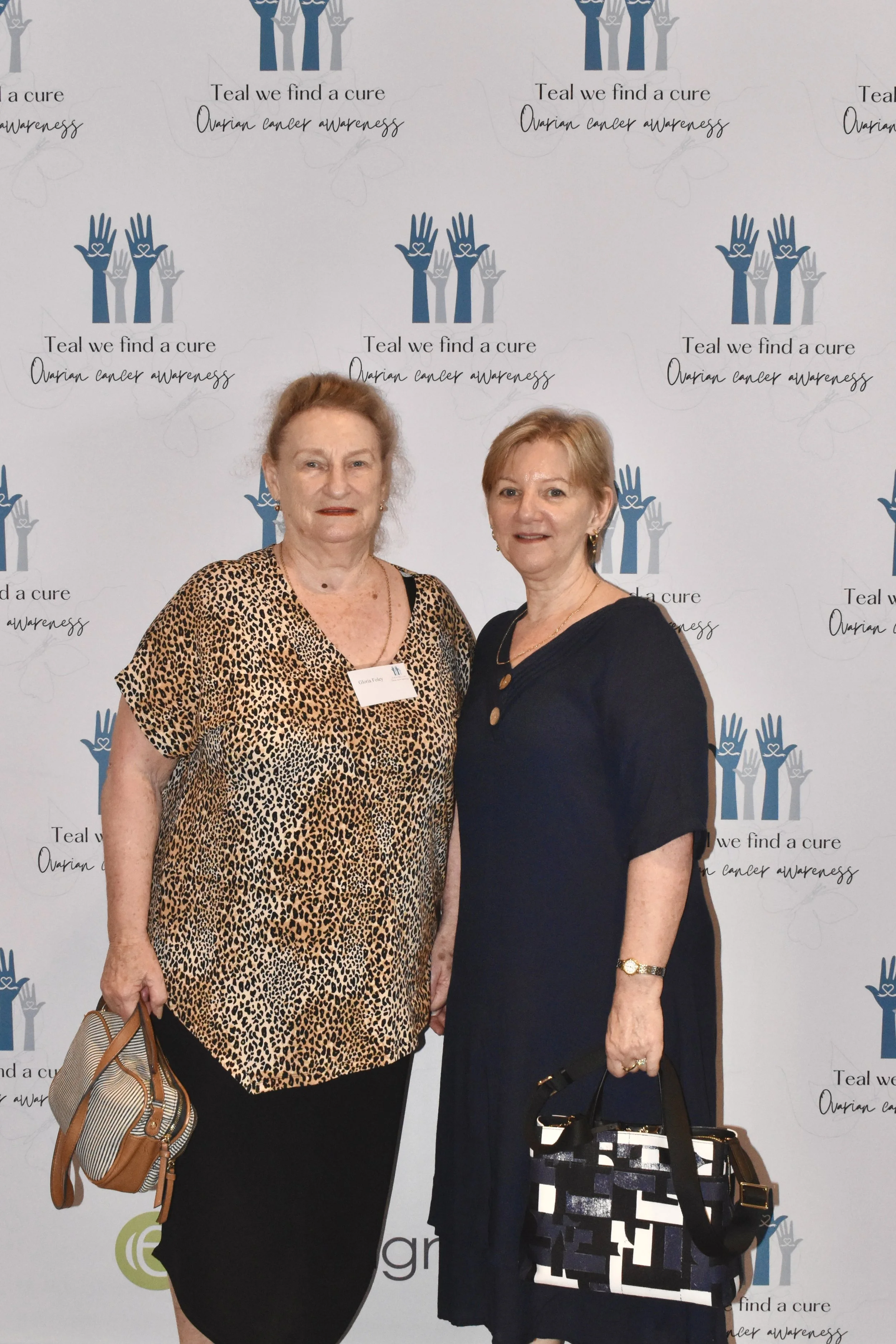 Two women standing in front of a backdrop with the logo and text for ovarian cancer awareness. One woman is wearing a leopard print top and black skirt, holding a tan purse. The other woman is wearing a navy dress and holding a black and white patter