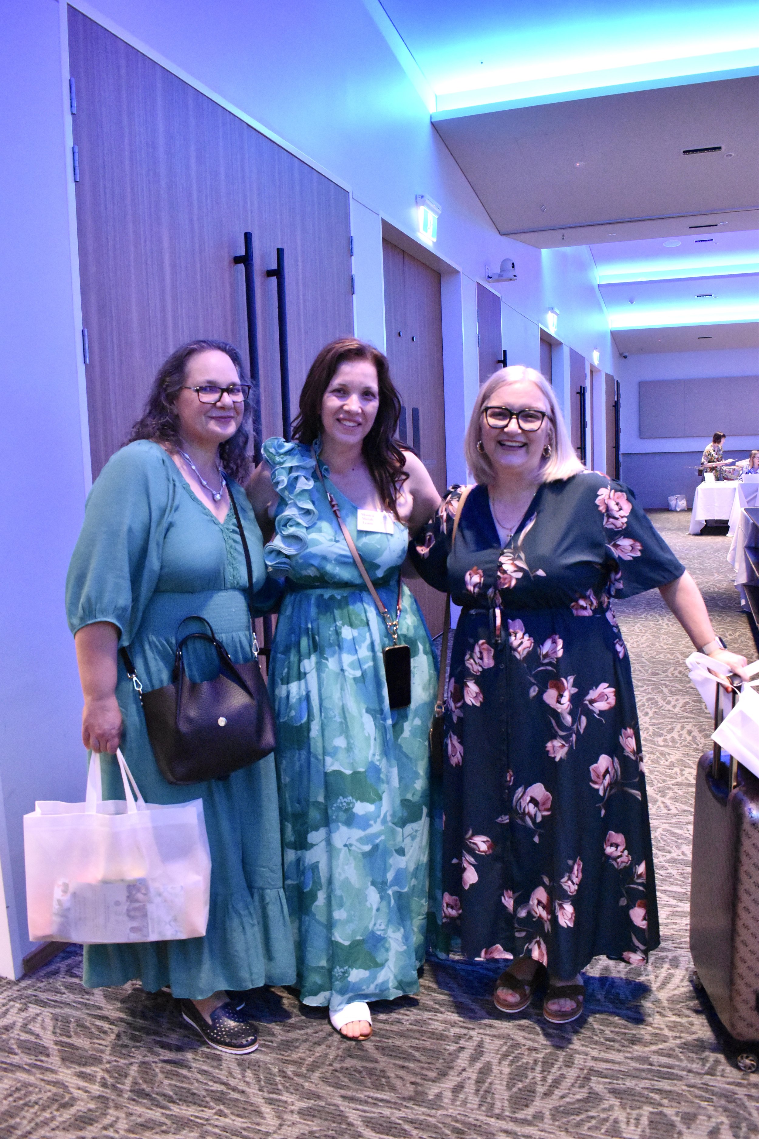 Three women smiling and posing together in a well-lit indoor conference or event space with wooden doors and tables in the background. The woman on the left is wearing glasses, a teal dress, and holding a tote bag. The woman in the middle has brown h